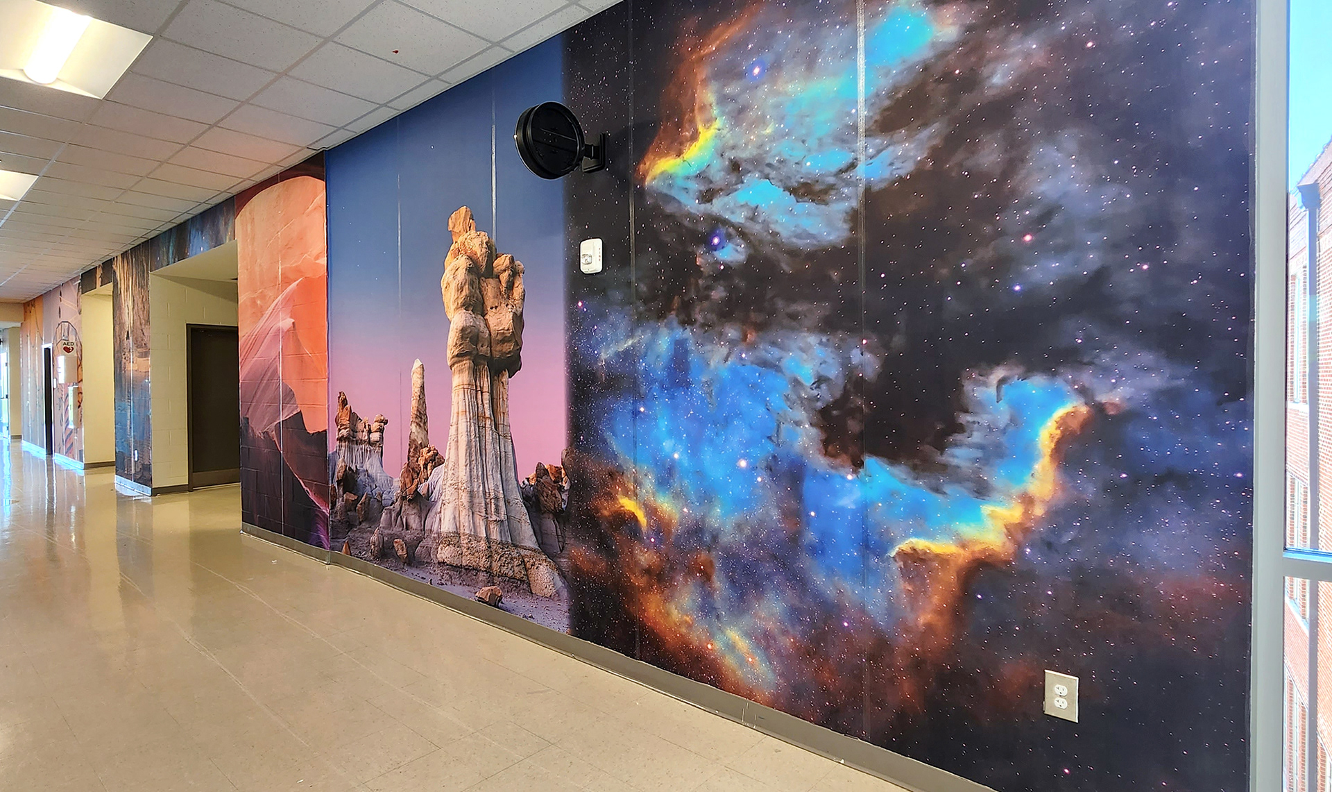 A look at the same floor-to-ceiling adhesive vinyl wall mural in a hallway view from the opposite angle. The mural features, from right to left, the image of galaxy, the image of colorful rock and sand formations, beyond that the images are too distant to decern clearly.