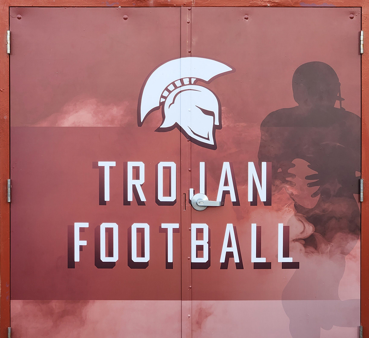 Full print adhesive vinyl door wrap with a burgundy background and white lettering that reads "TROJAN FOOTBALL" below a white image of a Trojan helmet. To the right of the door is a silhouette of a football player mid-play, holding a ball and running, partially visible behind the lettering.
