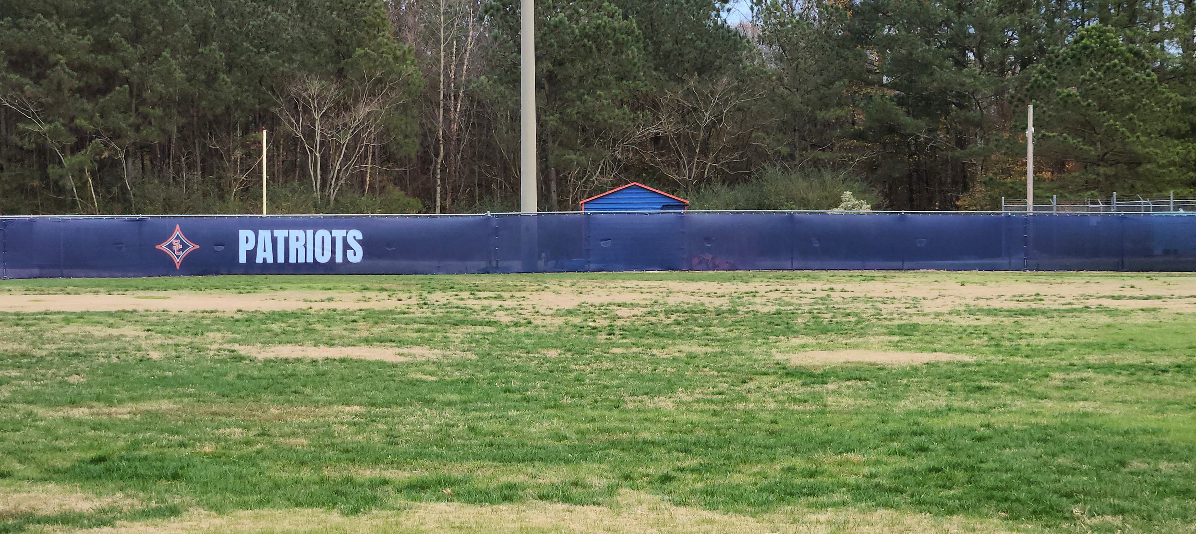 Navy blue fence windscreen installed on the outfield fence of a baseball field, featuring a small red SC logo at the center of a red four-point star with a white outline on the left, with the word 'Patriots' printed in white to the right of the logo.