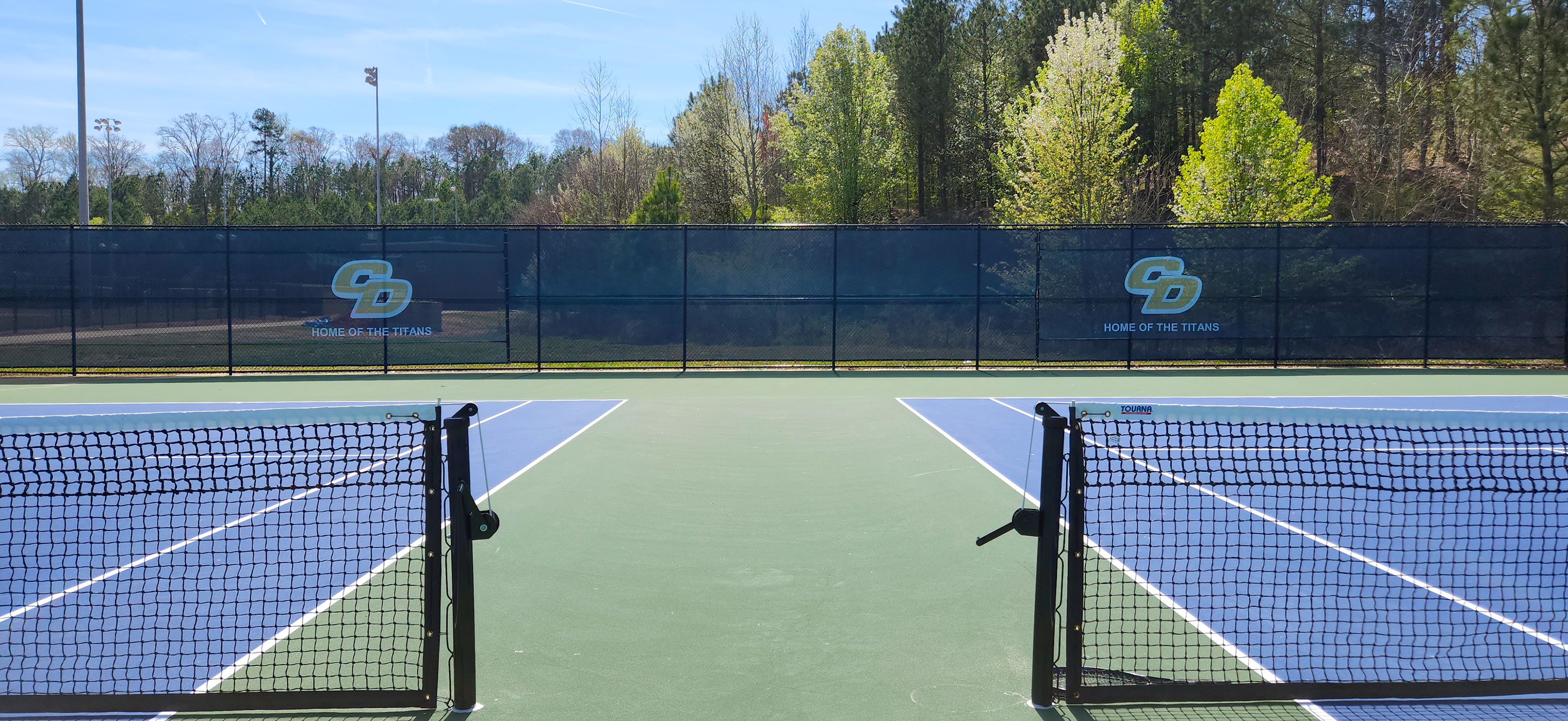 Blue fence windscreen surrounding a tennis court, featuring a yellow 'CD' monogram logo outlined in white, with the words 'Home of the Titans' displayed.