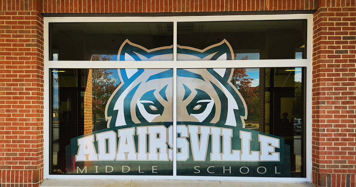 Brick building with a four-pane window featuring a large window perf decal. The decal shows the top part of a tiger's face above the word 'ADAIRSVILLE,' with the words 'MIDDLE SCHOOL' directly below.