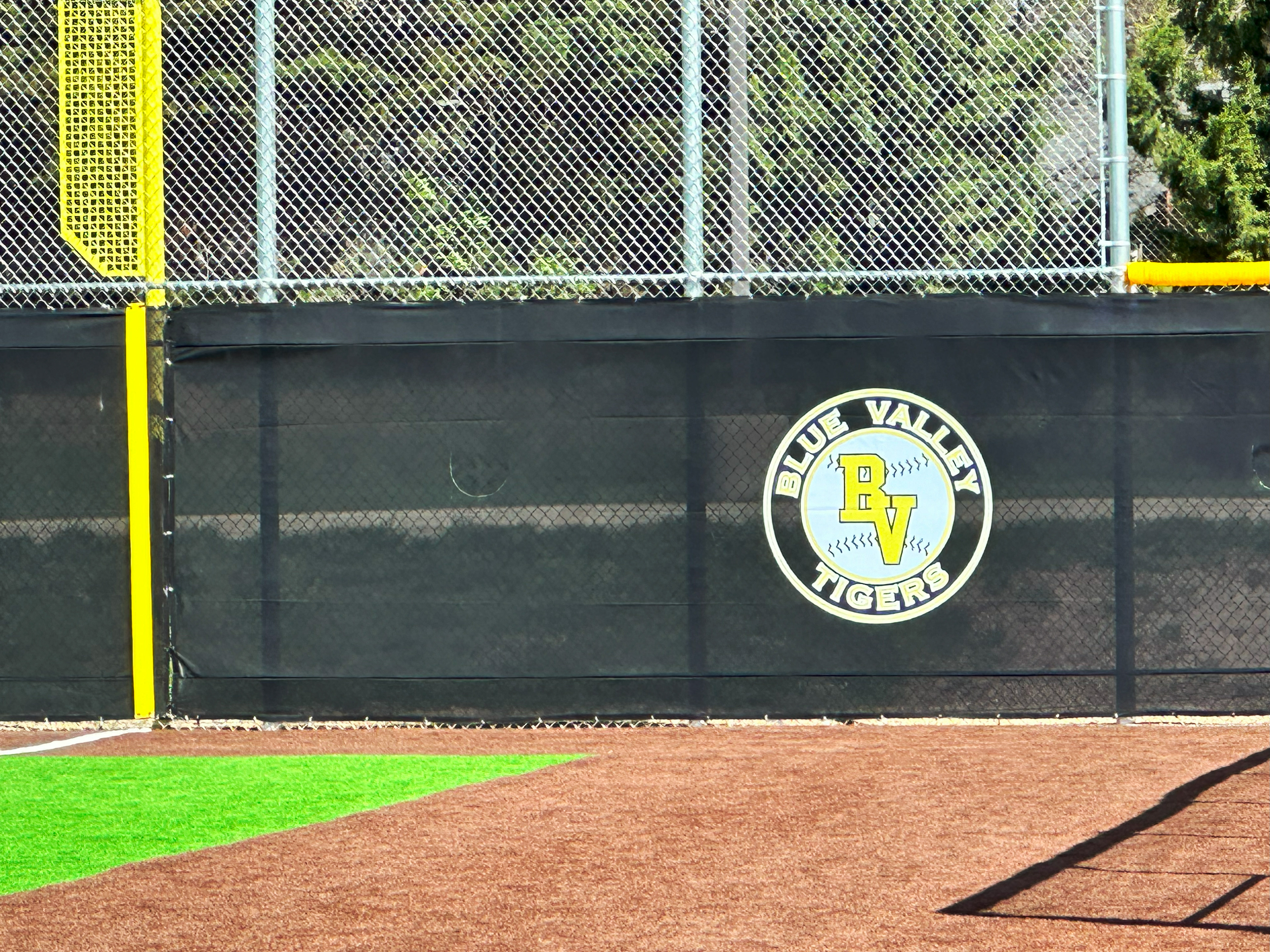 Closeup of black windscreen attached to a fence printed with a white and yellow round logo reading "Blue Valley Tigers" with a white baseball and the letter "BV" in the middle.