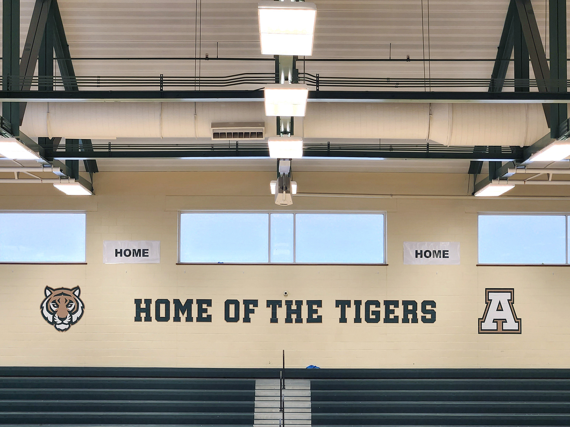 Adhesive vinyl decals on an off-white painted brick wall, placed below three windows and above a set of gym bleachers. From left to right: a tiger head logo, the words 'HOME OF THE TIGERS' in the middle, and an outlined monogram 'A' on the right.
