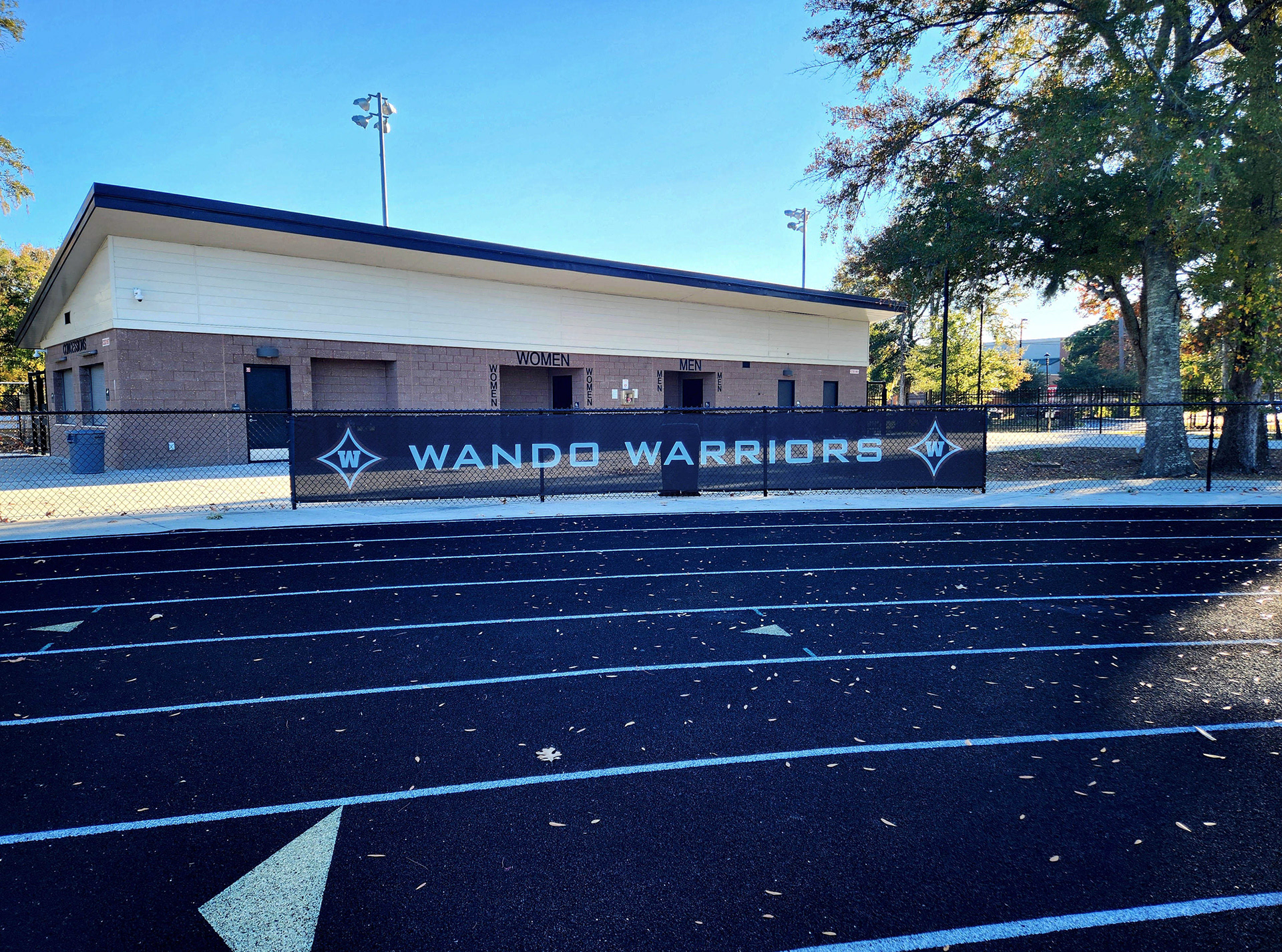 A black windscreen on a fence near a field. The windscreen features the text 'Wando Baseball' in white, with the 'W' logos on both sides. Each 'W' logo consists of a large navy four-point star outlined in white and red, with a white 'W' in the center of the star.