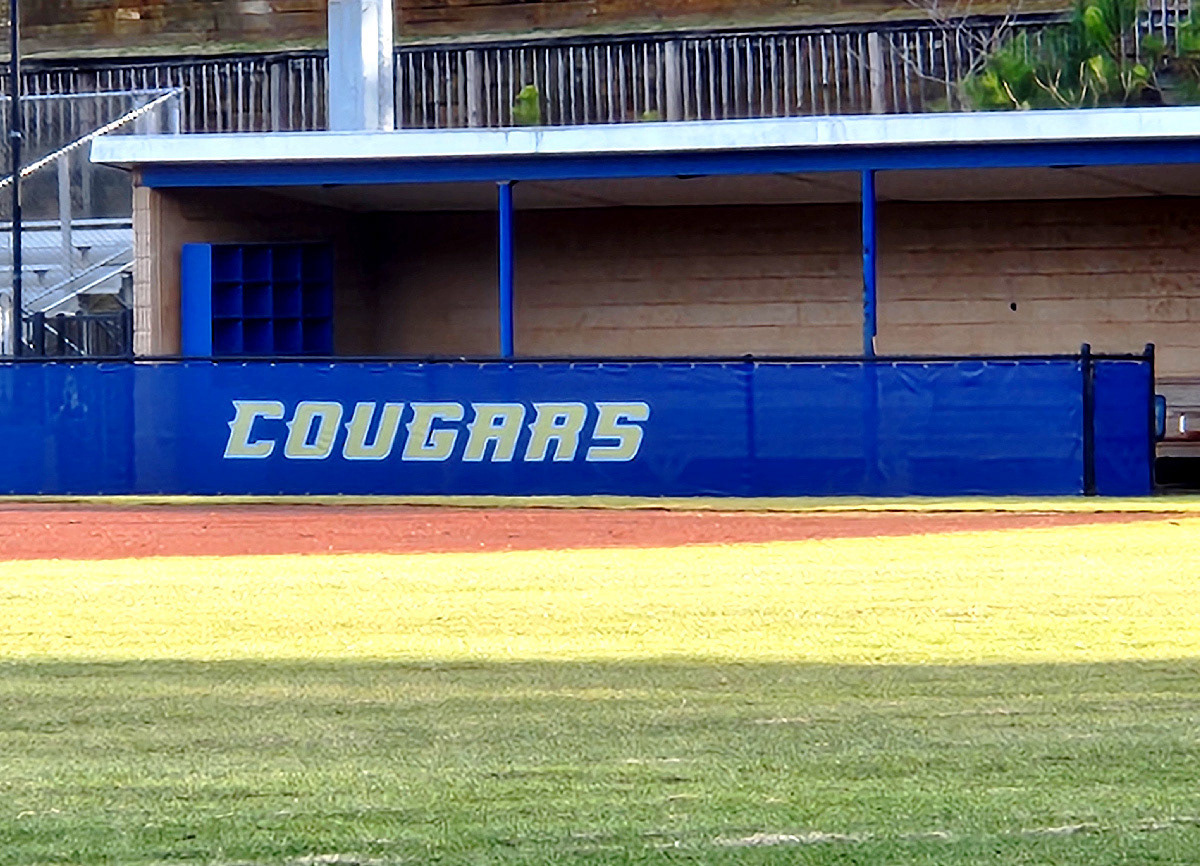 blue windscreen printed with yellow text outlined in white reading 'cougars' attached to a fence barrier surrounding a baseball dugout
