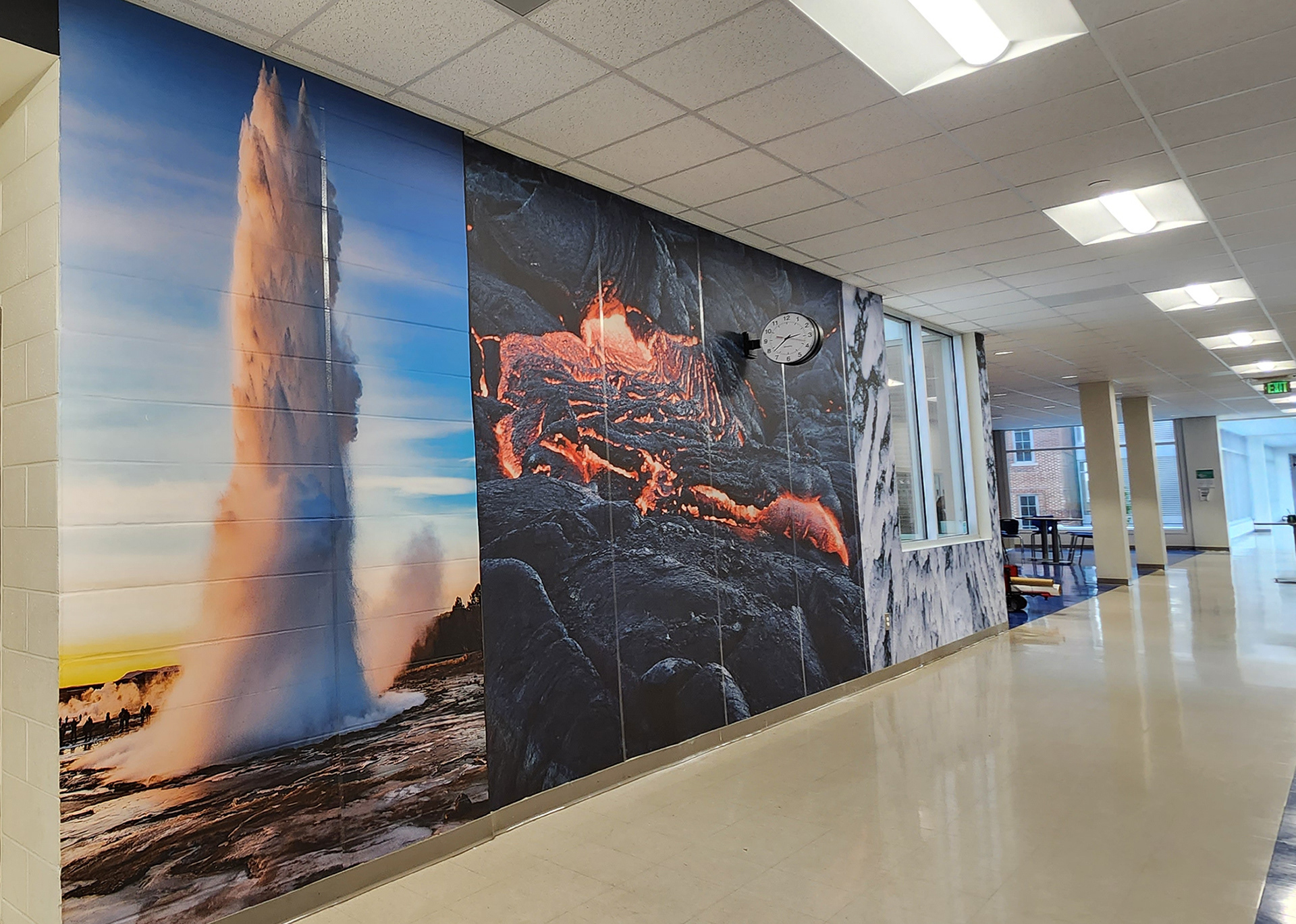 A floor-to-ceiling adhesive vinyl wall mural in a hallway. The mural features, from left to right, a geyser, lava among a volcanic cliffside, a glacier, and beyond that, an extending hallway with windows. 