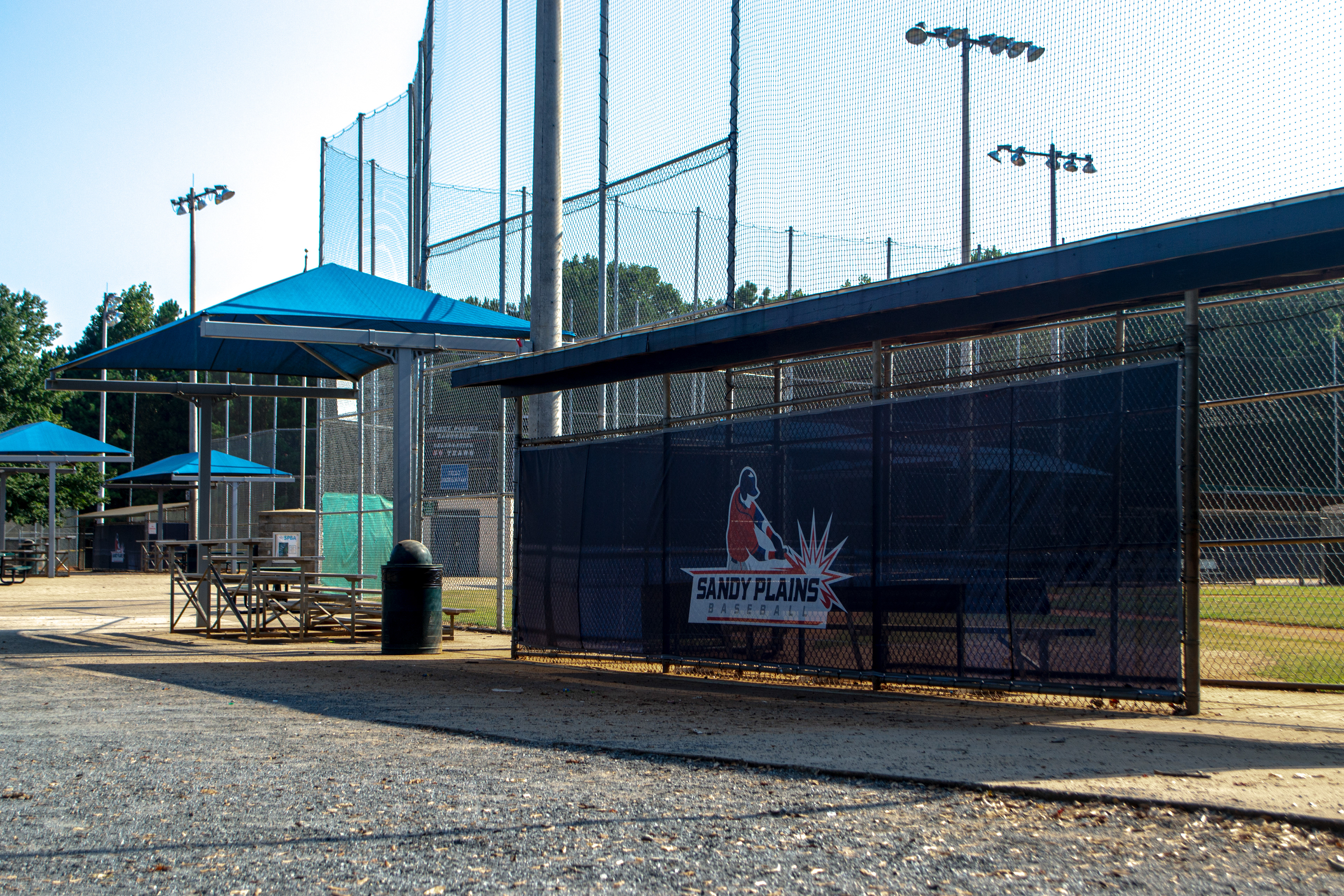 A dugout viewed from a nearby walkway, located next to a baseball field. The dugout has a large branded windscreen panel at the back. The panel shows an image of a baseball player in a red shirt and navy hat hitting a ball, outlined in white. Below the image, the words 'SANDY PLAINS BASEBALL' appear in navy blue on a white background, with two horizontal red lines above and below the text.