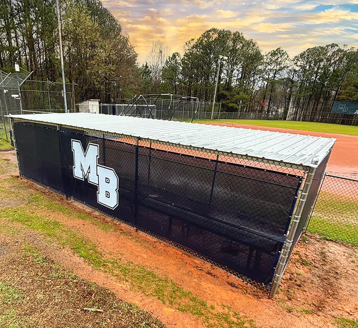 Navy blue fence windscreen branded with "MB" in white, wrapped around a baseball dugout, viewed from above from the back right side.