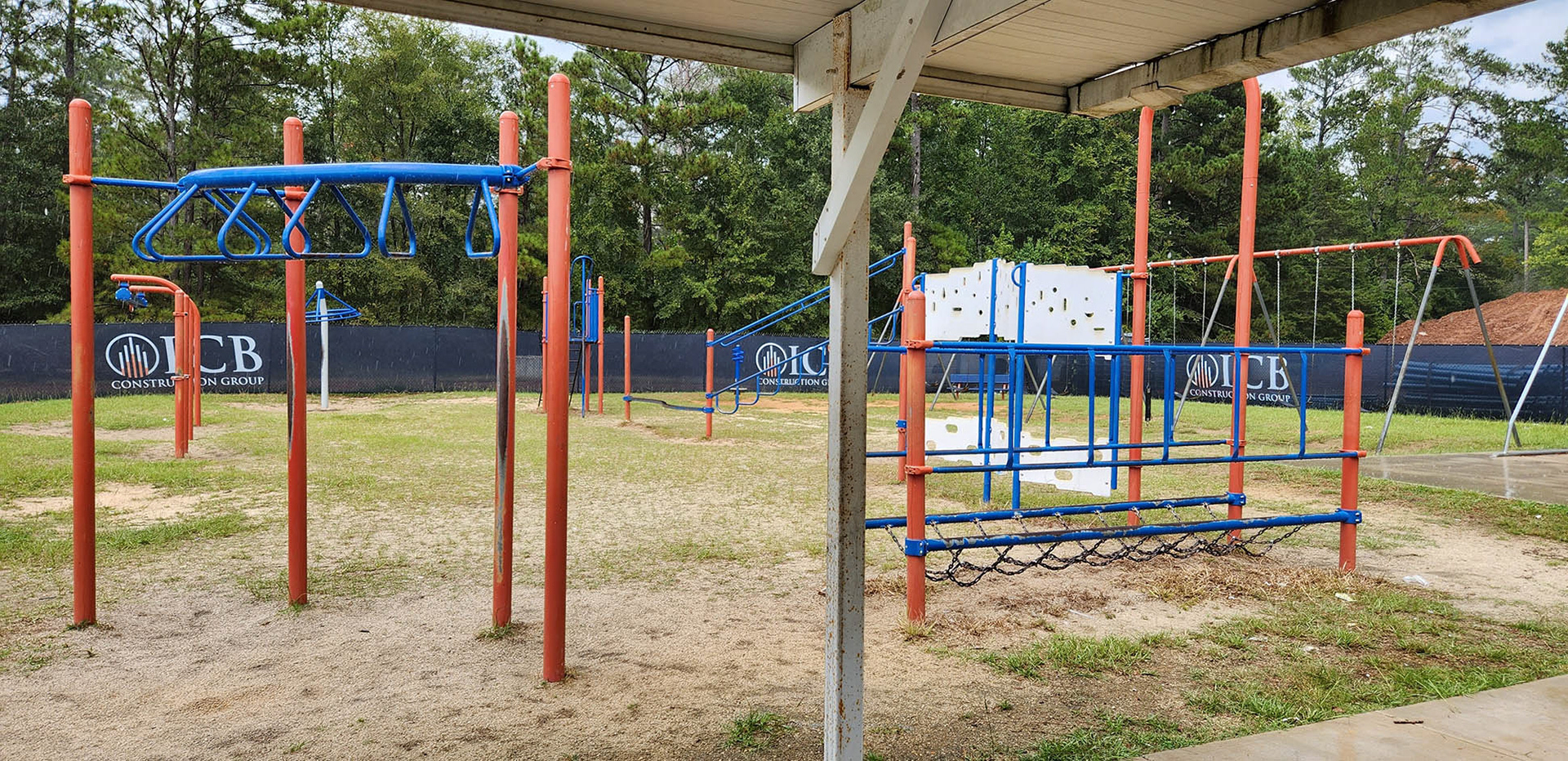 A navy windscreen separating a playground and a construction site. The windscreen has three logos printed on it, each consisting of a circular white outline with five orange and white lines of varying lengths arranged from bottom to top. Next to each logo, the text 'ICB' is printed in white, and beneath the logo, the text 'CONSTRUCTION GROUP' is also printed in white. 