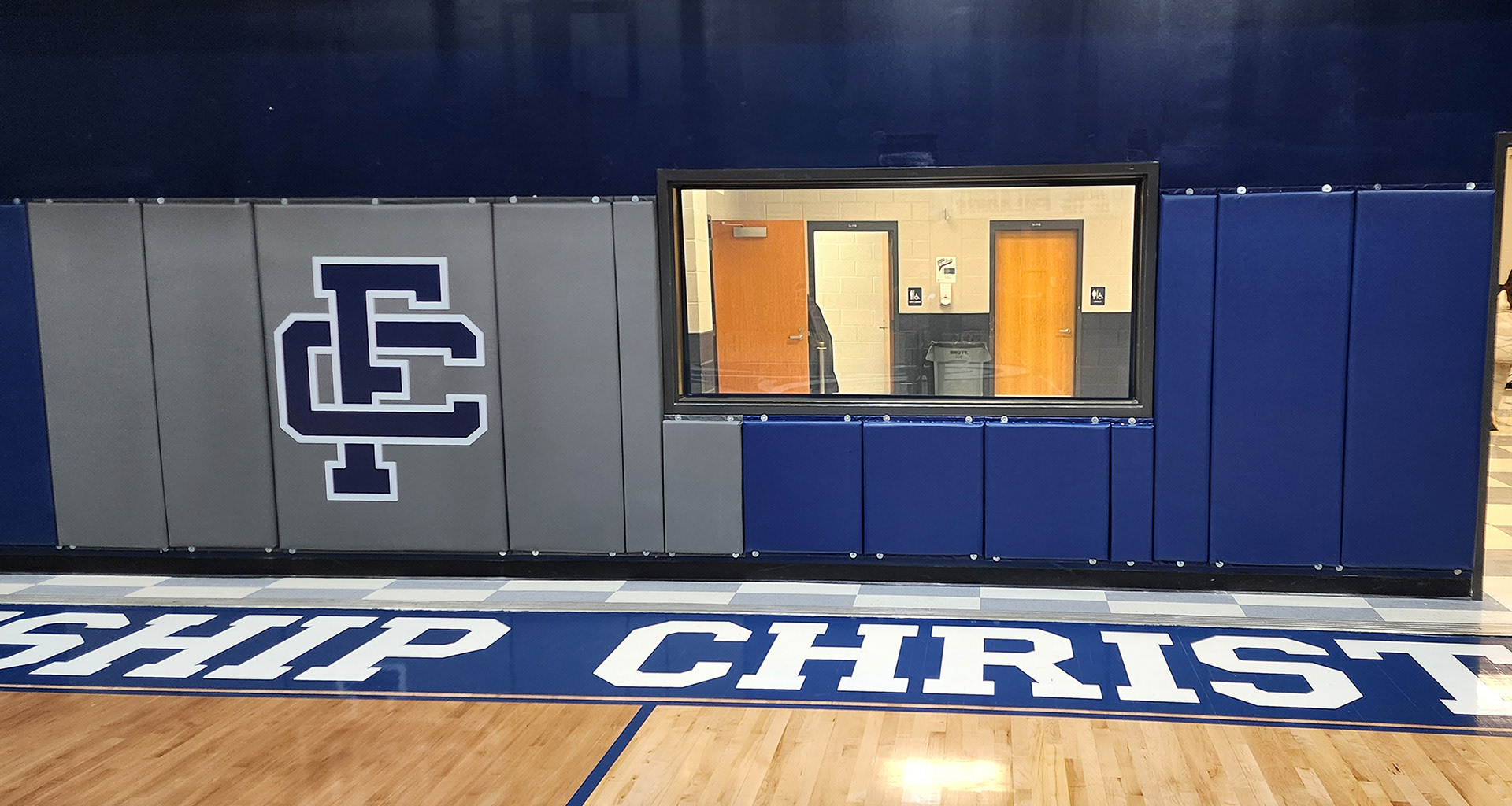 Fourteen vinyl-covered backstop pads in a gym, attached to a wall below and beside a large window. The left pads are gray, with one pad featuring a navy blue 'FC' monogram logo outlined in white. The pads on the right are blue.