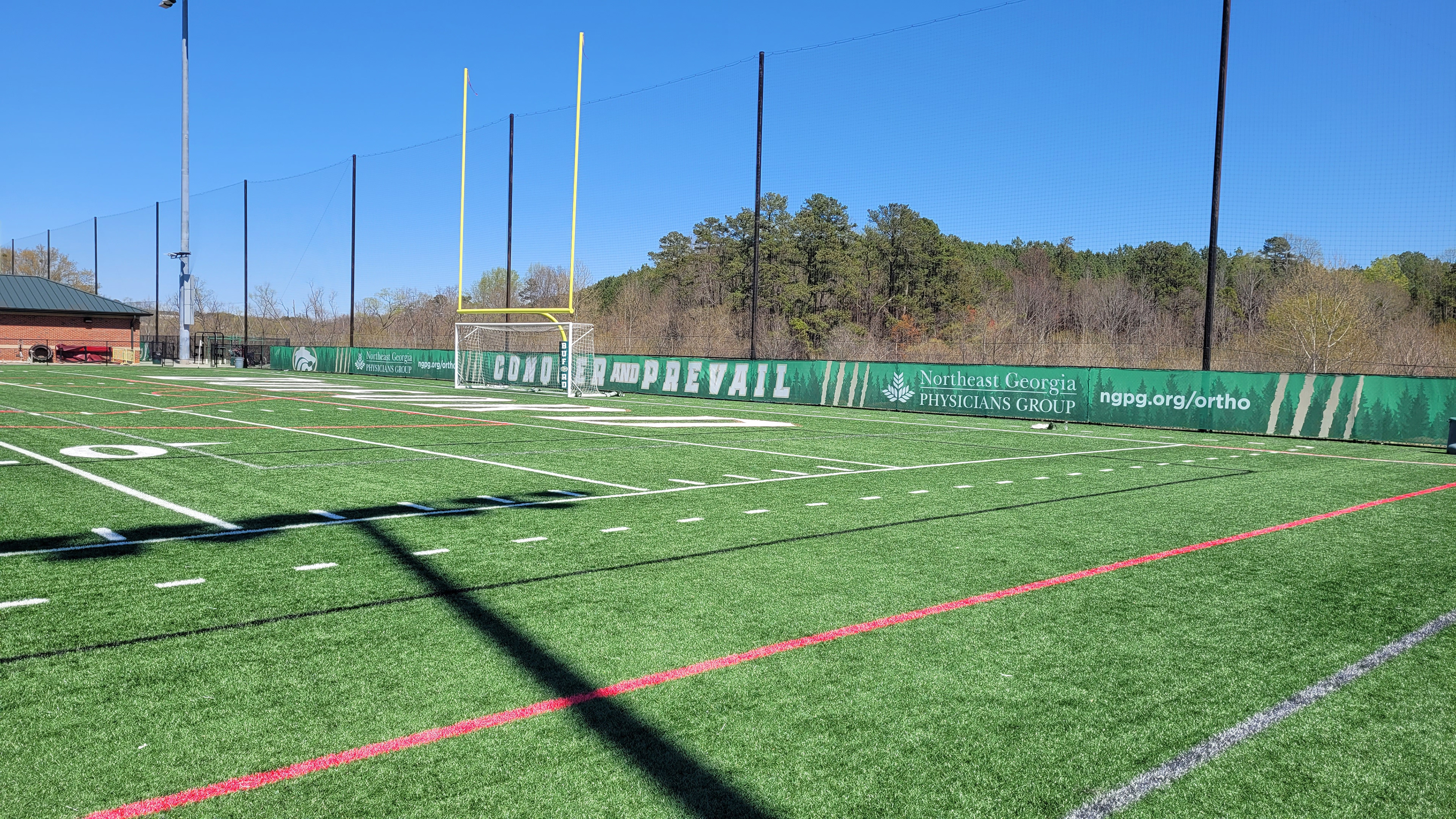 Green fence windscreen branded with a dark evergreen tree silhouette, white lettering with a beige outline reading "CONQUER AND PREVAIL," and beige slashes next to sponsor messages, at the end of a football field. A football goal post with protective foam padding covered in branded green sports vinyl at the base, with black sports netting placed above the fence directly behind the goal for safety.