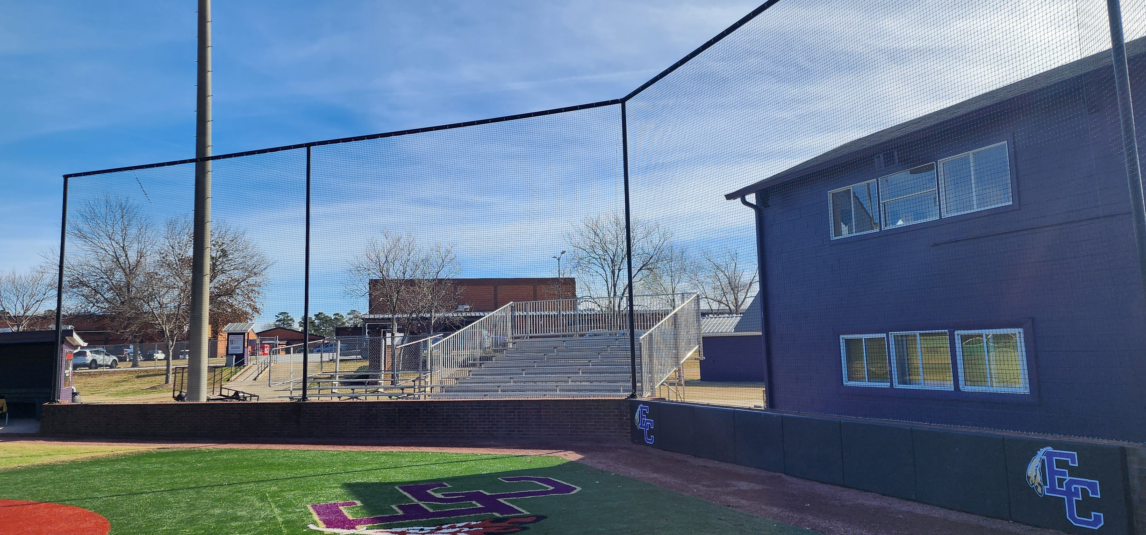 backstop black sports netting surrounding the catcher's box above black sports vinyl covered backstop padding printed with blue 'EC' logo with white outline and white and yellow feather accents, attached to a brick wall