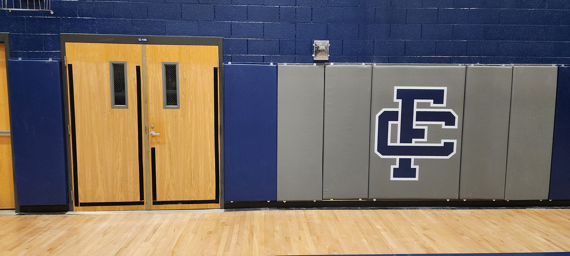 Eight vinyl-covered backstop pads, with two blue pads on the left and a double door in the middle. The pads on the right are gray, with the middle gray pad featuring a navy 'FC' monogram logo outlined in white.