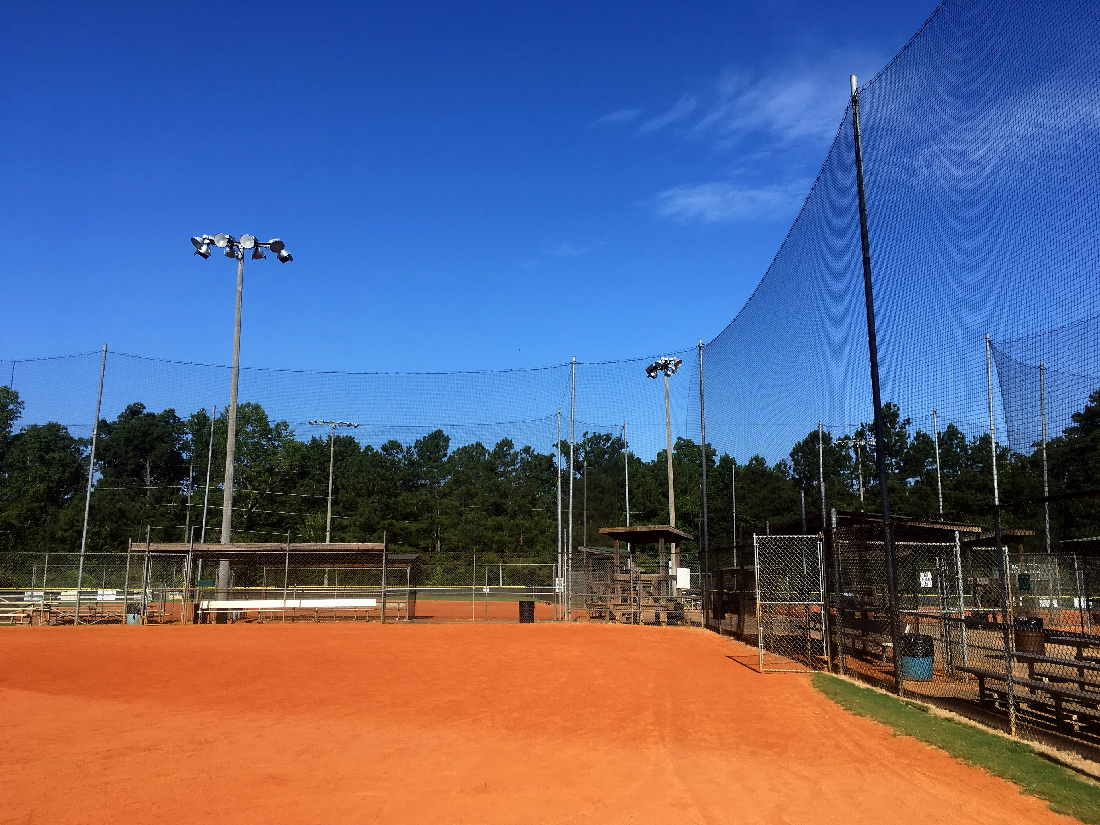 A view from inside the baseball field shows vertical sideline netting surrounding the field to the front and right. Also to the front and right, there is a set of metal bleachers with a nearby baseball dugout, both located behind the netting. In the distance, another baseball field is partially visible.