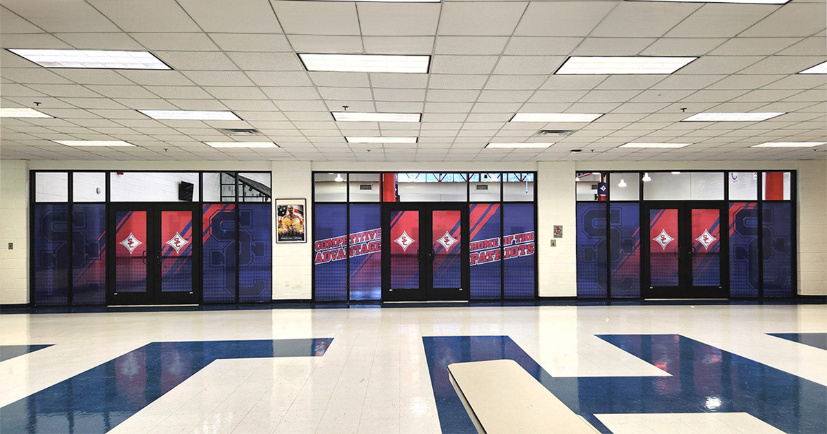 A red, white, and blue window perforation featuring the SC logo, with the text 'Competitive Advantage' and 'Home of the Patriots' displayed on three glass doors inside a school
