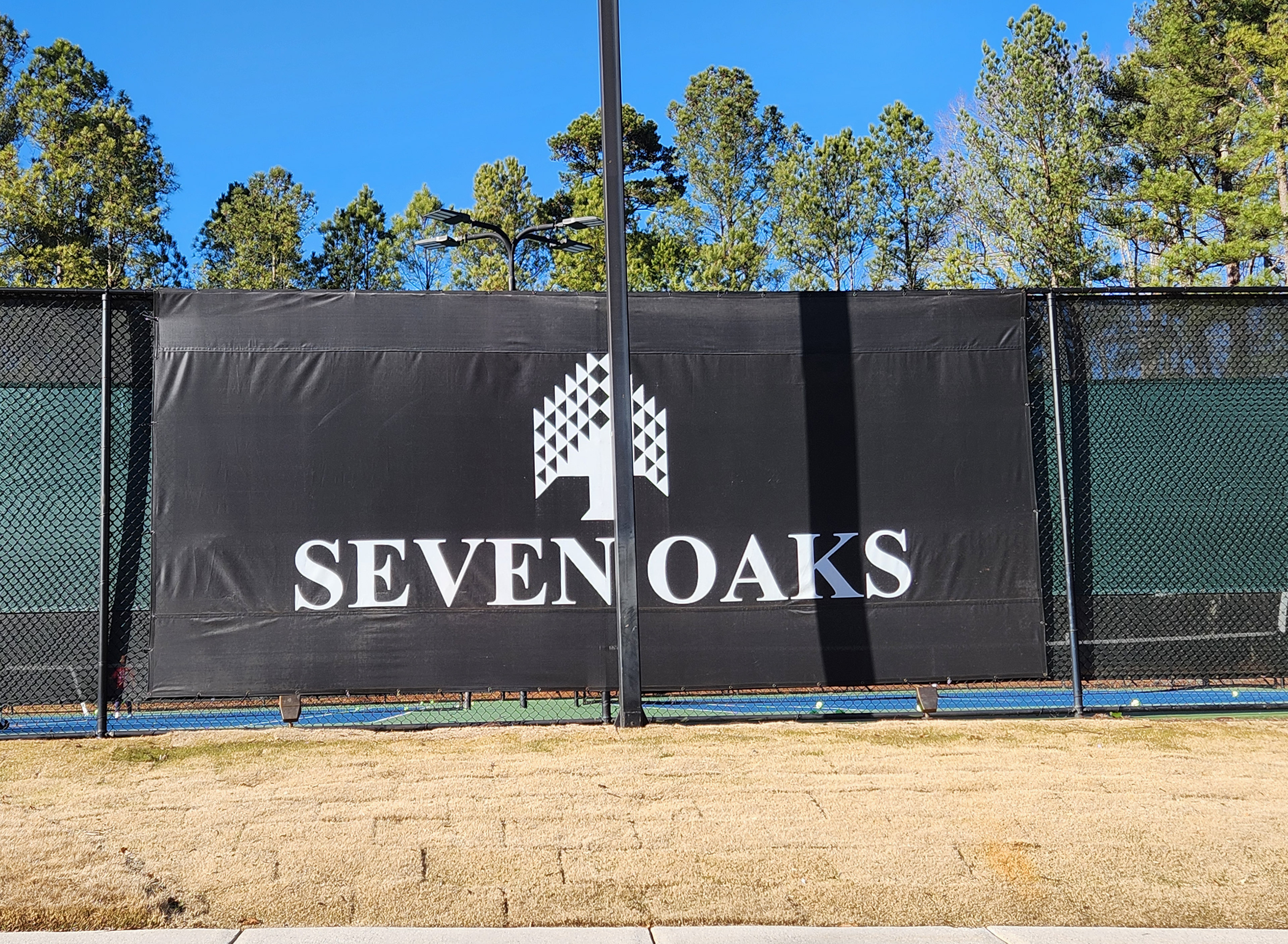 green fence windscreen bordering a tennis court, overlayed with a black windscreen banner in the middle branded with white geometric tree logo and text reading 'seven oaks'