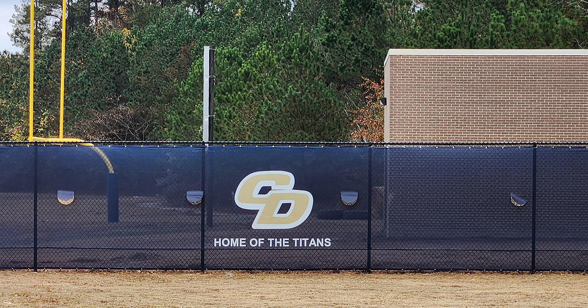 Close-up of a blue fence windscreen around a baseball field, showing a 'CD' monogram logo in beige outlined in white, with the words 'Home of the Titans' beneath the logo.