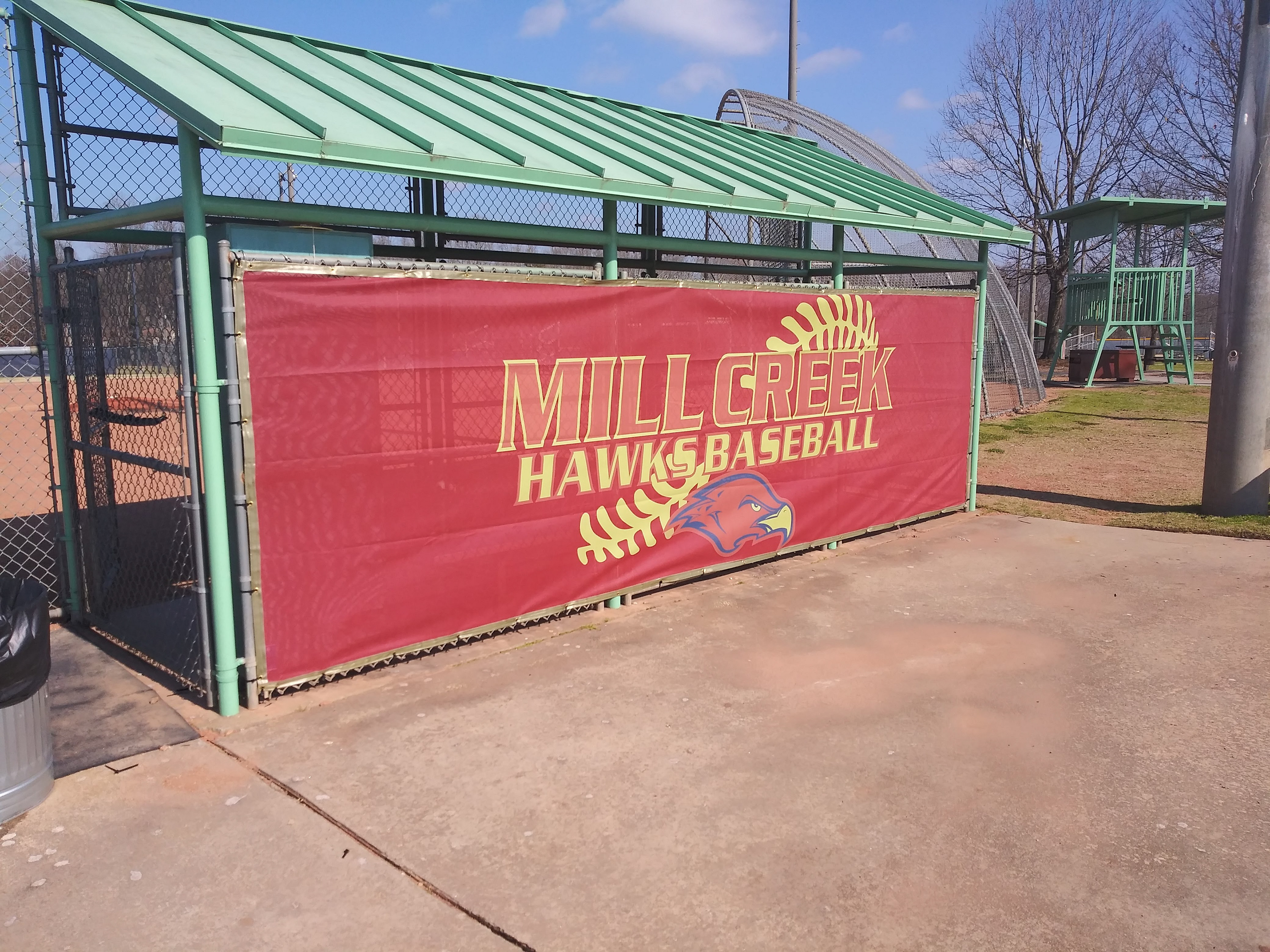 A fenced in baseball dugout with a red windscreen panel with gold binding, and printed with the words "MILL CREEK" in a lighter red outlined in yellow, and the words "HAWKS BASEBALL" in yellow oulined in a lighter red. Behind the text printed in yellow are the laces of a baseball, below that head of a red bird with a yellow beak outlined in blue.