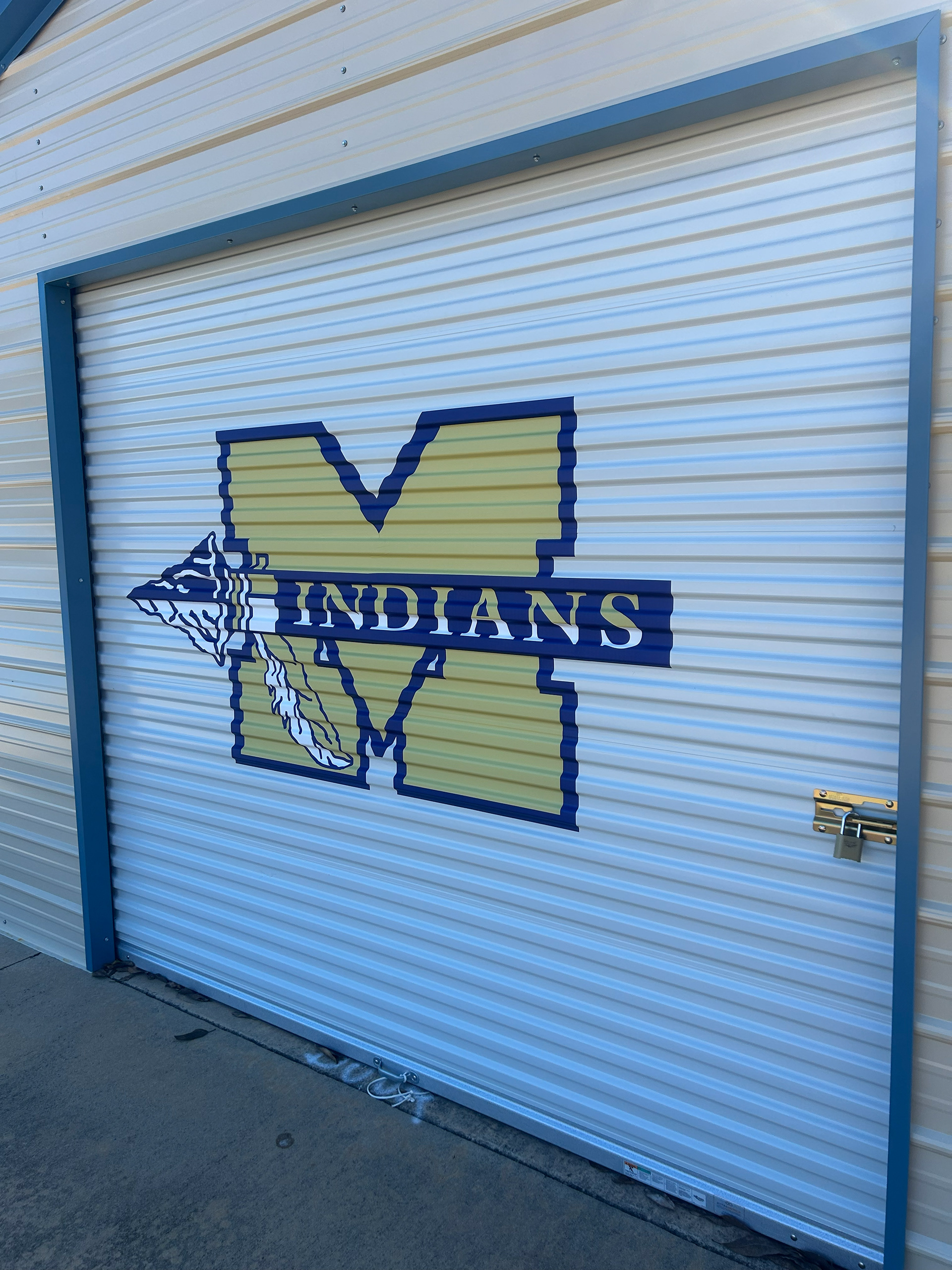 Close-up side view of a large custom-designed vinyl sports team logo adhered to a metal rolling garage-style door on a barn-style school building.