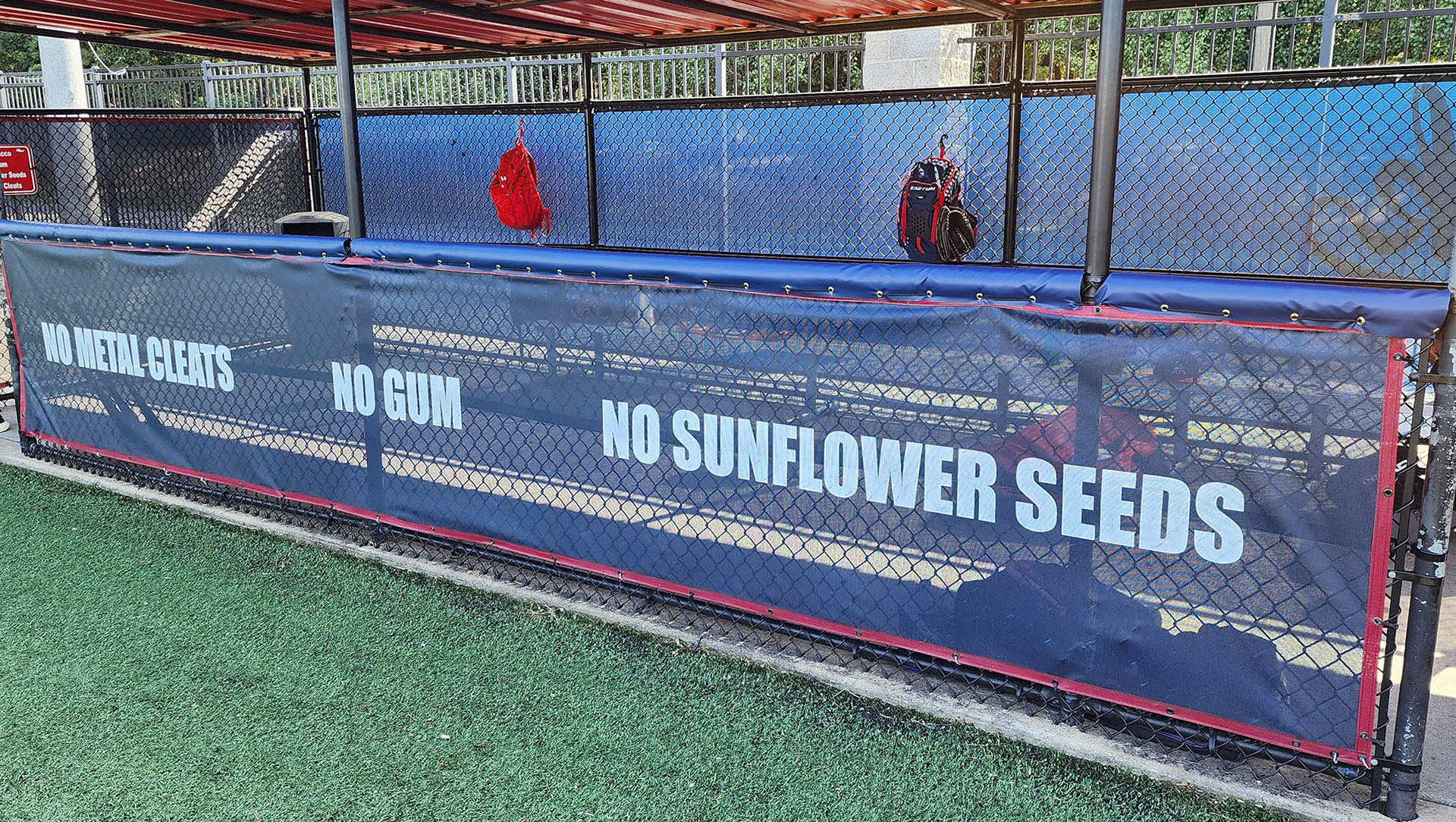 A fenced-in baseball dugout with blue windscreen panels affixed to the front, back, and sides. The front panel has red binding and is printed with the words 'NO METAL CLEATS,' 'NO GUM,' and 'NO SUNFLOWER SEEDS' in white text, with red binding around the panel. Attached to the top of the front dugout railing is blue vinyl-covered rail padding. 
