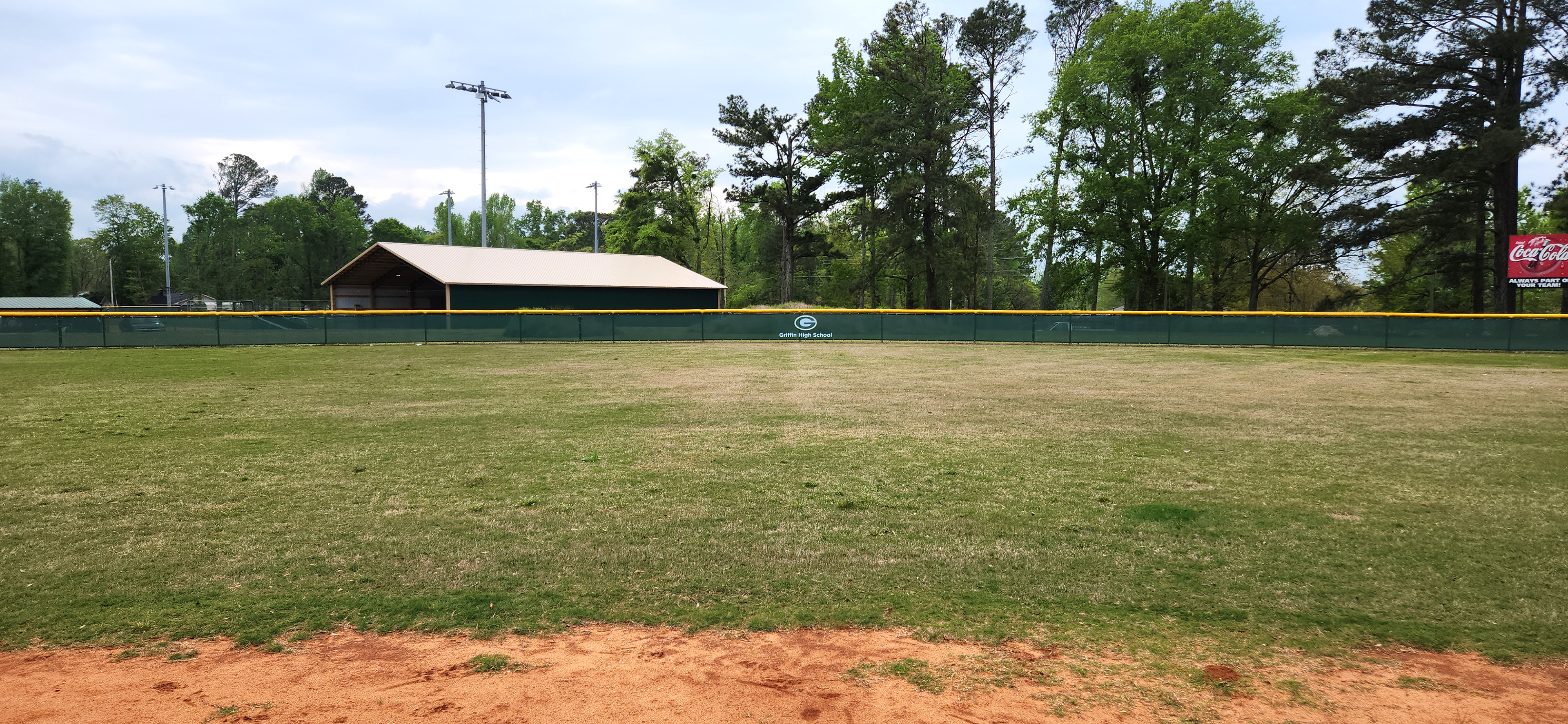 Distance shot of dark forest green windscreens are branded with a white and green 'G' logo and white text reading 'Griffin High School' attached to a fence with a yellow corrugated poly cap rail top fence cap surrounding a baseball field.