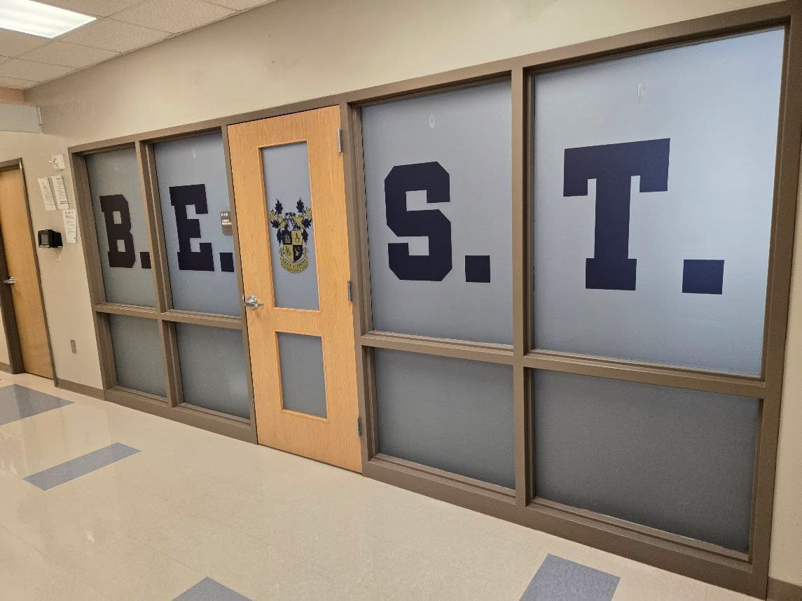 Right-angled view of an interior school doorway featuring a windowed door and surrounding windows with perforated graphics. Navy blue letters and a centered yellow and navy blue school crest are visible across the top windows, with frosted glass filling the remaining space.