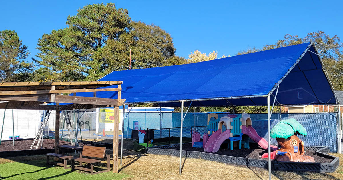 A play area beneath a large, metal-framed blue shade structure shaped like a pavilion, next to a seating area covered with a wooden shade structure.