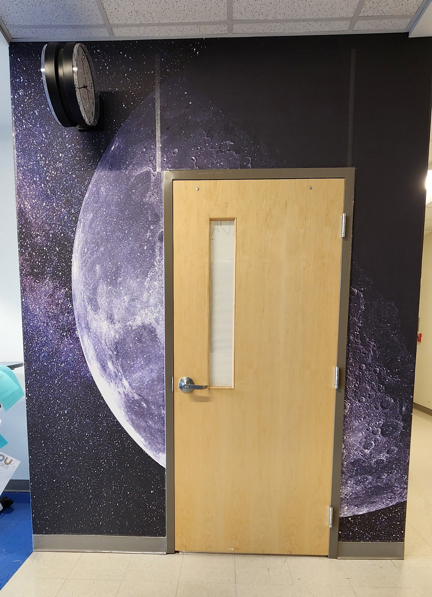 A floor-to-ceiling adhesive vinyl wall mural in a hallway, surrounding a closed door. The mural features a purple-tinted moon with space filled with stars in the background. 