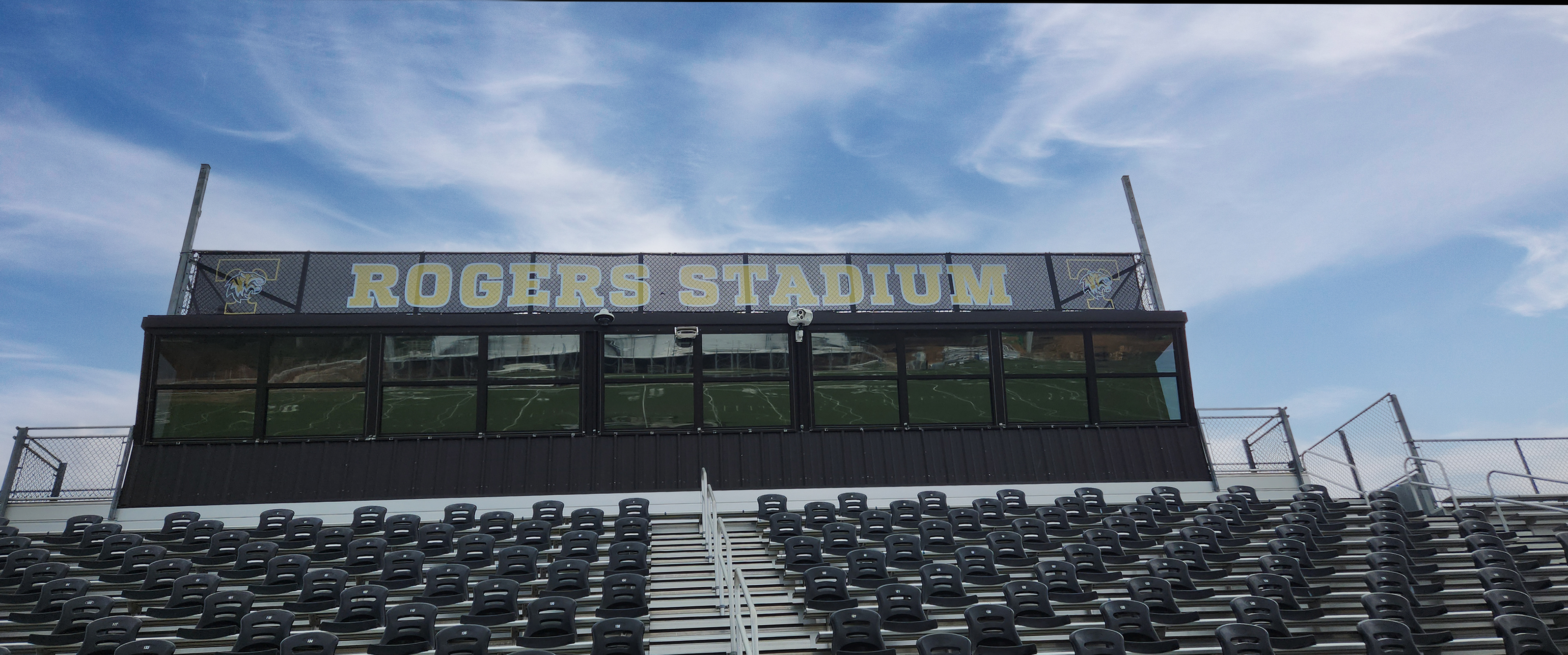 Black windscreen branded with the 'tiger nation' logo and the words "Rogers Stadium" printed in yellow with a white outline attached to fencing a top a press box above bleacher seating.