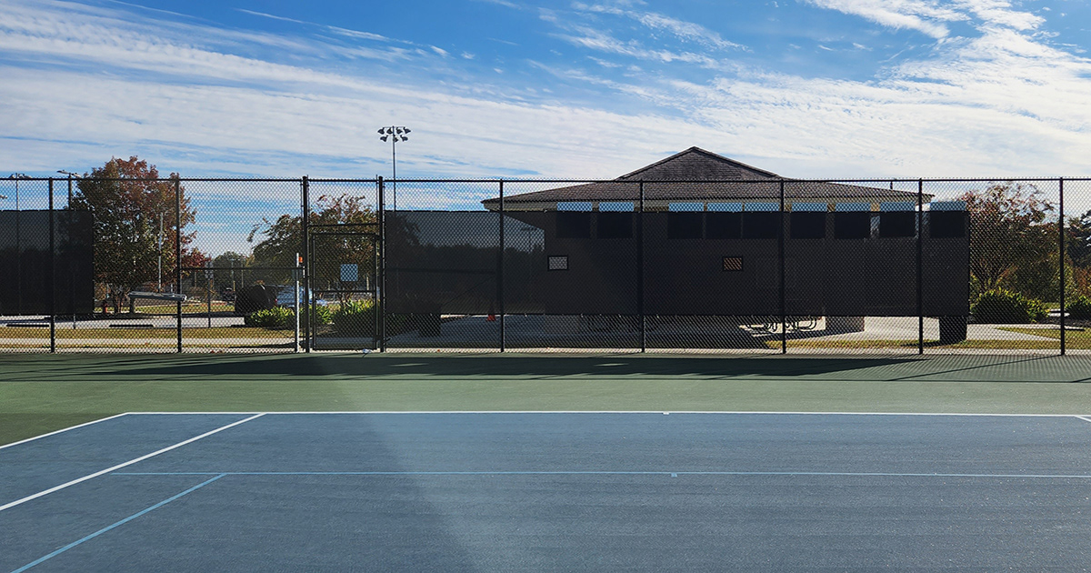 Tennis courts with a fence enclosure, affixed with two black windscreen panels featuring square vents.