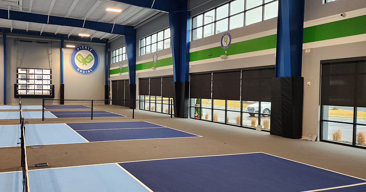 Black vinyl-covered padding over metal support beams surrounding four indoor pickleball courts, and the bottom of a protruding window trim.
