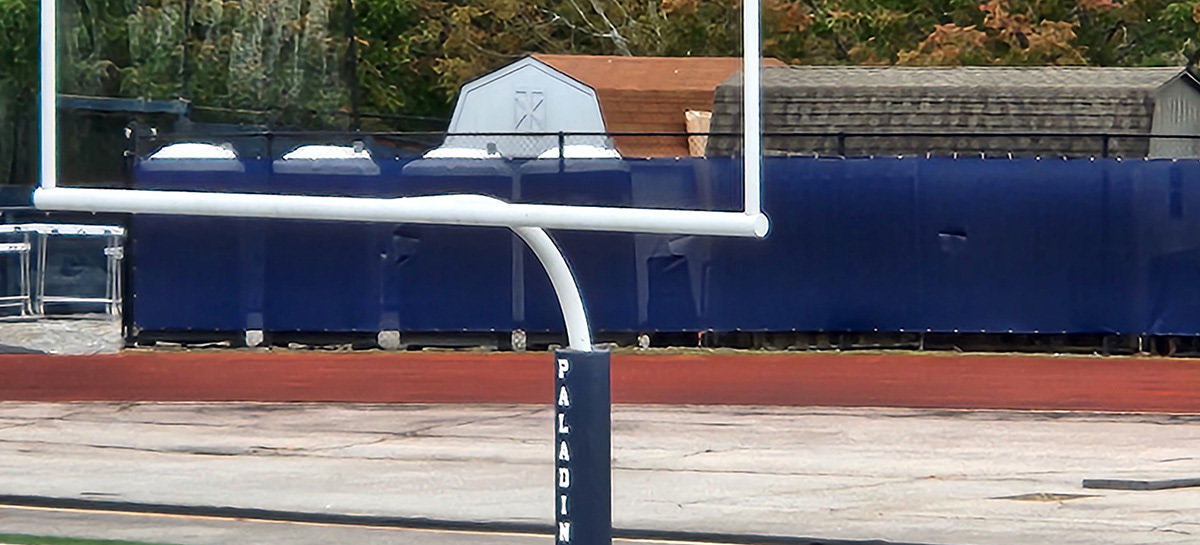 A Navy blue fence windscreen is positioned behind the goal, separating the football field from two buildings and four porta potties just outside the fenced area.