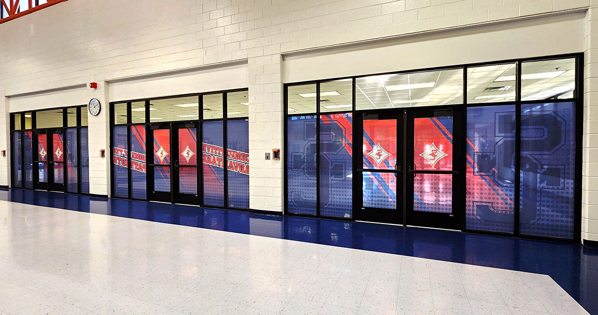 A red, white, and blue window perforation featuring the SC logo, with the text 'Competitive Advantage' and 'Home of the Patriots' displayed on three glass doors inside a school