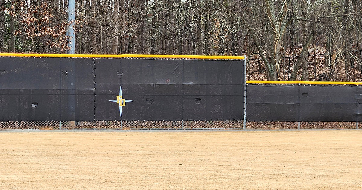 black fence windscreen printed with a black four point star outlined in white under yellow lettering with a white outline reading 'FD'', affixed to a fence around a baseball field, topped with a yellow fence guard rail top tear drop fence cap
