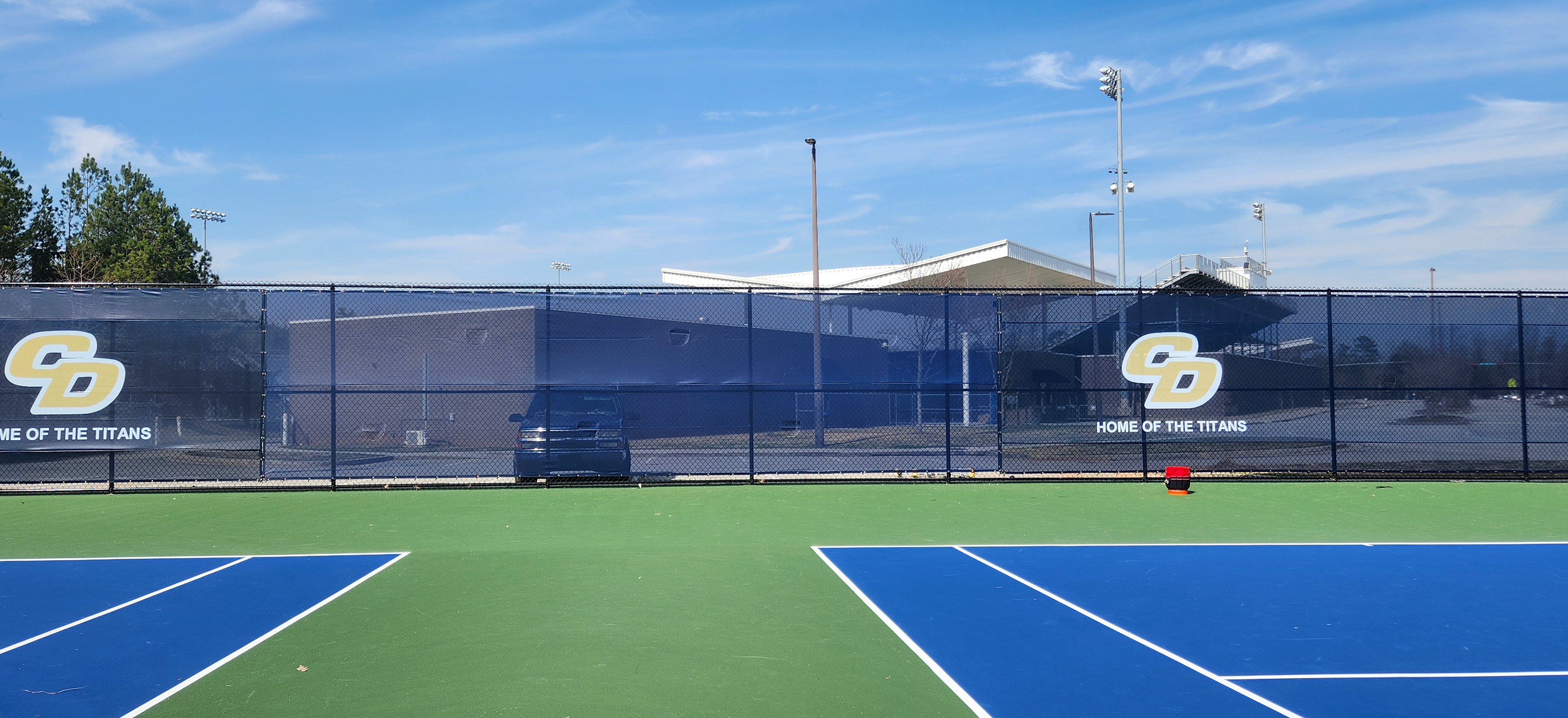 Blue fence windscreen surrounding a tennis court, featuring a yellow 'CD' monogram logo outlined in white, with the words 'Home of the Titans' displayed.
