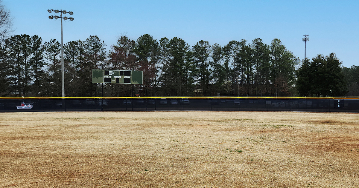 A view of a baseball field's fence on a sunny day, with trees and a building in the background. The fence has a yellow corrugated poly-cap rail top and a black branded windscreen attached. The windscreen displays a logos to the left and a number to the right several feet away. The logo shows a baseball player in a red shirt and navy hat hitting a ball, outlined in white, with the words 'SANDY PLAINS BASEBALL' in navy blue on a white background, and red lines above and below the text. The numer is "229" displayed vertically printed in white .