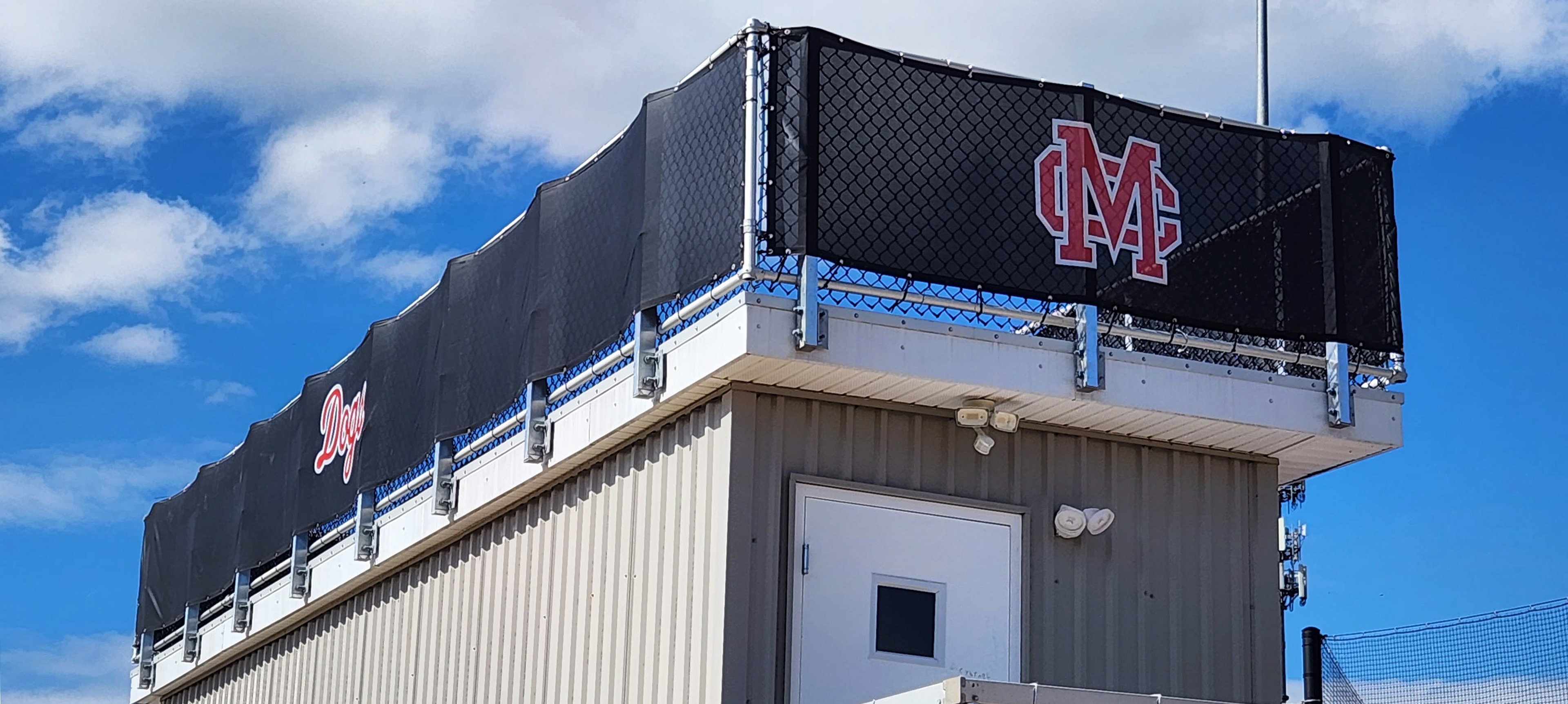 Branded black windscreen affixed to fencing, wrapping around the top of a press box. One side is printed with the word 'Dogs' in red outlined in white, while the other side, over a door, displays a red 'MC' monogram logo outlined in white..