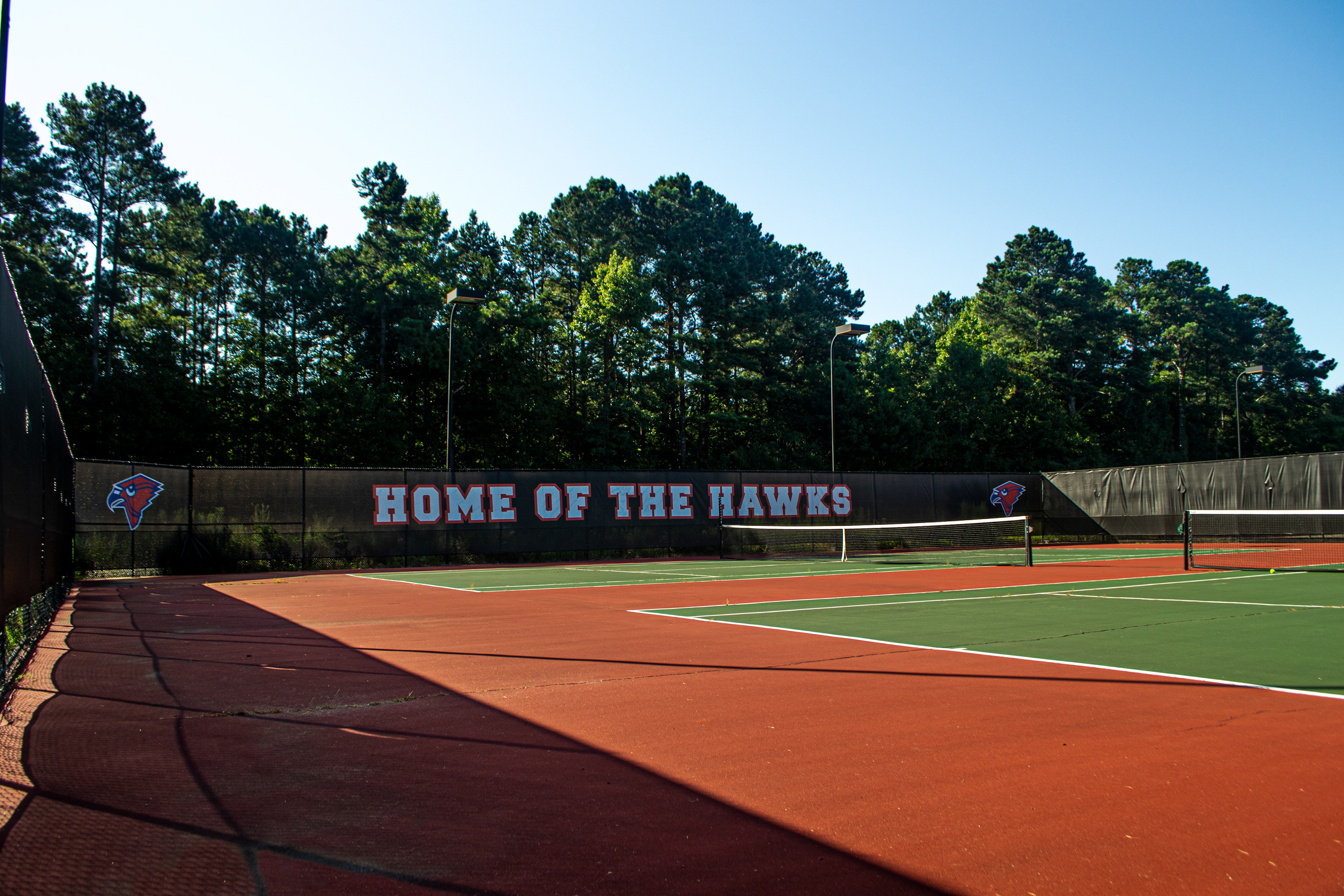 Branded fence windscreen panels surrounding a tennis court. At the far end, a windscreen displays the text 'HOME OF THE HAWKS' in white letters outlined in red. To the left and right of the text are red cardinal head logos outlined in white.