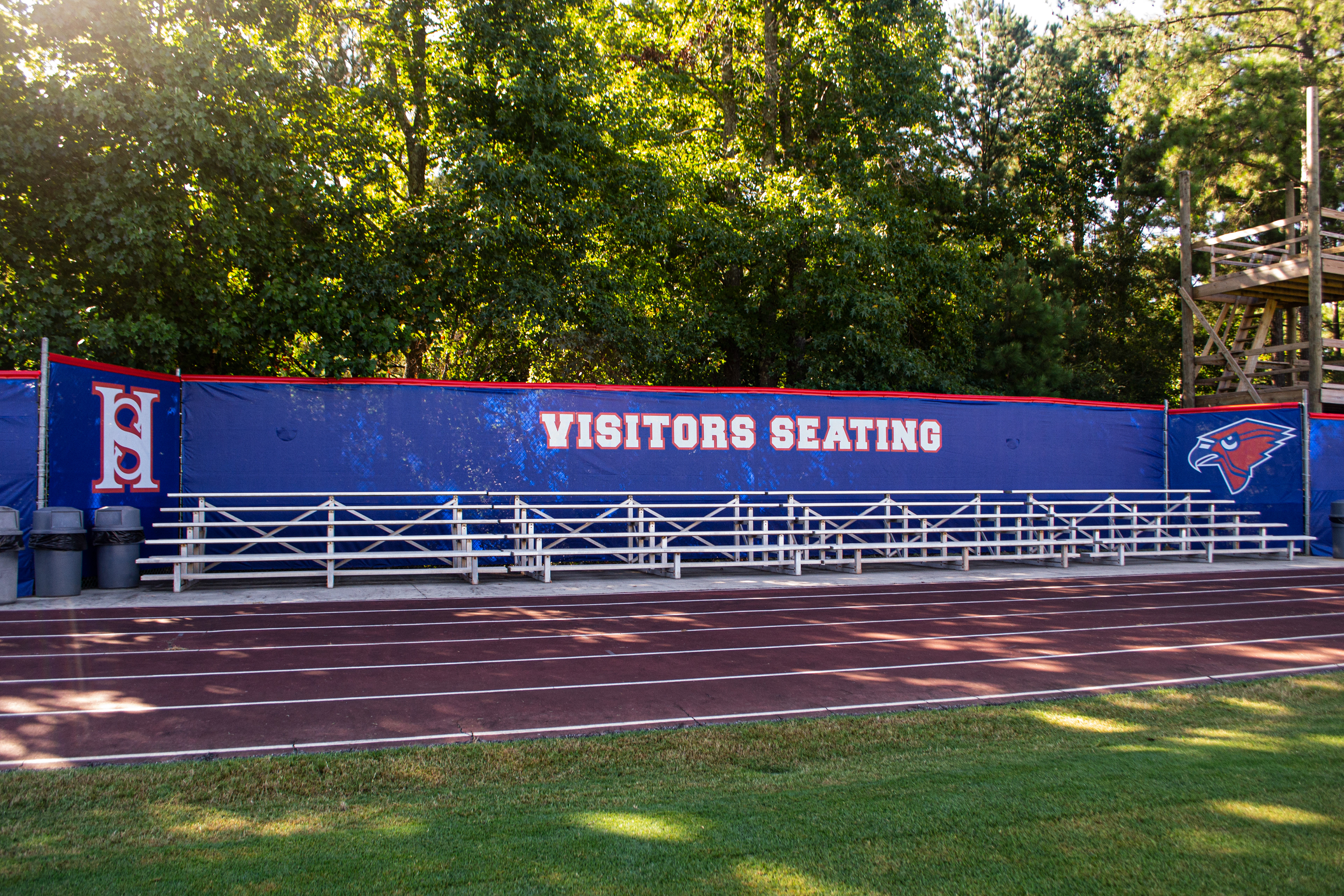 In the foreground, grass and a running track are visible, followed by metal bleachers backed by a fence with a branded windscreen and red fence cap, with trees in the background. The windscreen, from left to right, features a white 'HS' monogram logo outlined in red, the words 'VISITORS SEATING' in white outlined in red, and a red cardinal head outlined in white.