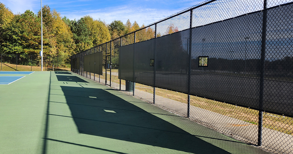 Tennis courts with a fence enclosure on the right, featuring three black windscreen panels with square vents affixed to the fence.