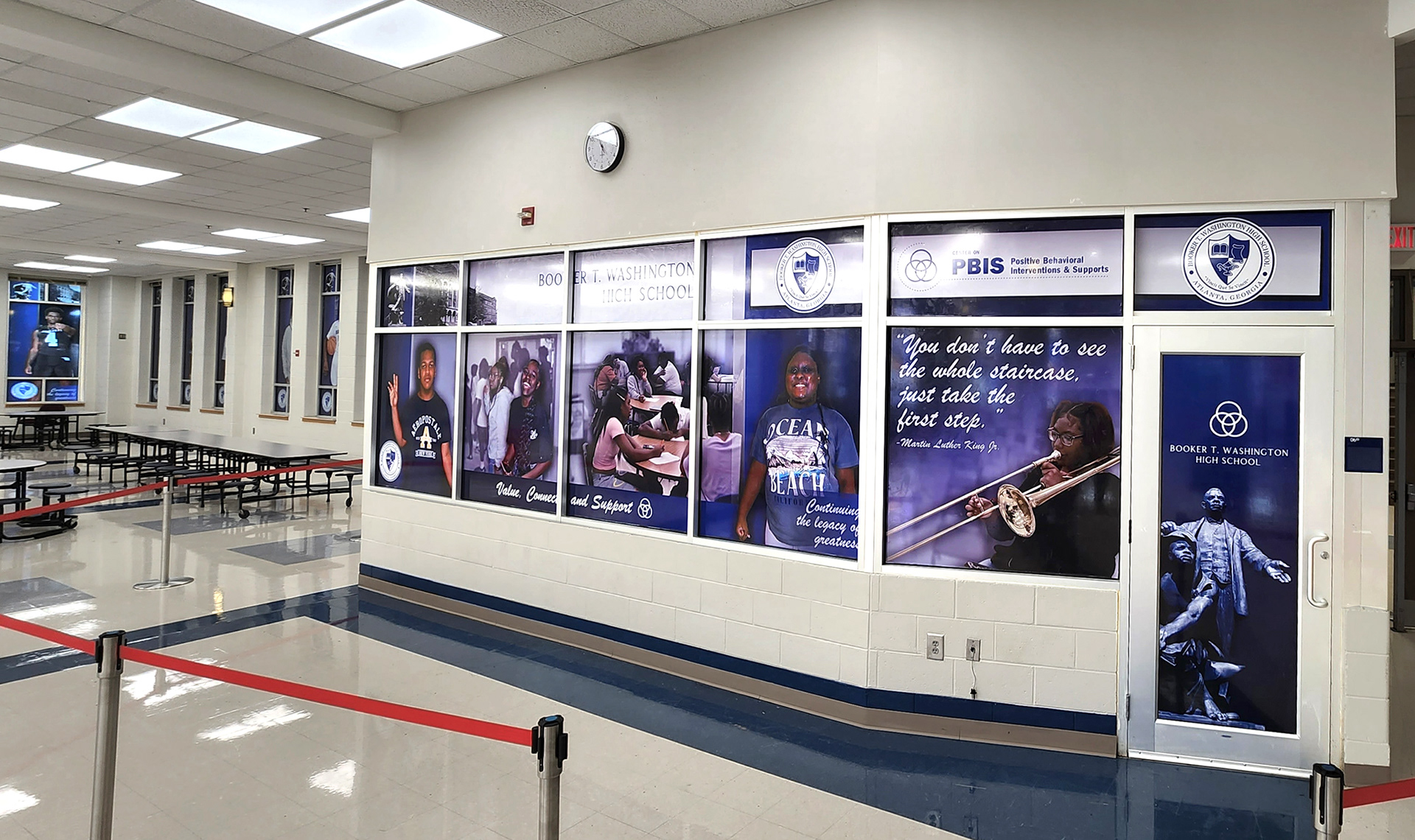An office inside a high school cafeteria, surrounded by windows. The windows display several images of students, along with motivational phrases, the name of the school, various logos, and an image of the statue of Booker T. Washington. In the background, within the cafeteria, additional images of student athletes are displayed on the windows. All images are created using full print window perf.