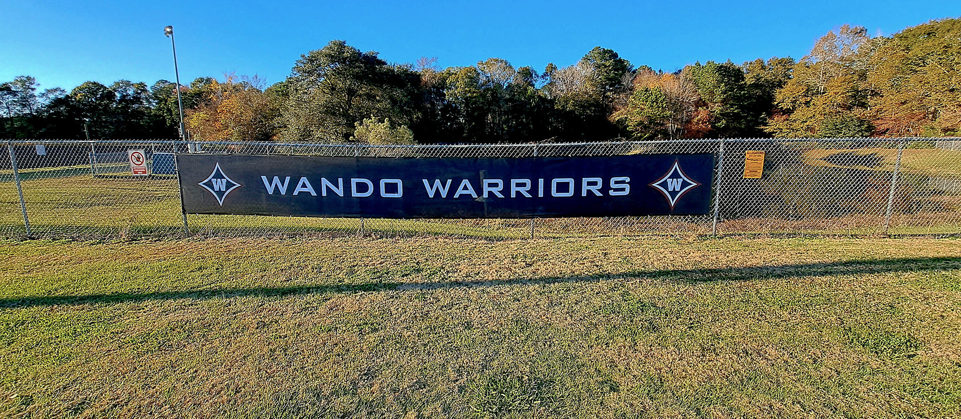 A black windscreen on a fence near a field. The windscreen features the text 'Wando Baseball' in white, with the 'W' logos on both sides. Each 'W' logo consists of a large navy four-point star outlined in white and red, with a white 'W' in the center of the star.