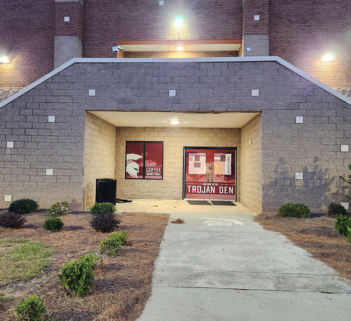 The entrance to a school building, with two doors on the right and a double-pane window on the left. The doors are covered with full-print adhesive vinyl, and the windows at the center top of the doors are covered with window perf. Together, the vinyl and window perf create an image of two darker burgundy Trojan helmet silhouettes facing each other, with a Trojan metal shield in the middle. A dark burgundy banner beneath the shield reads 'WELCOME TO THE TROJAN DEN' in white text, with a burgundy smoke background.