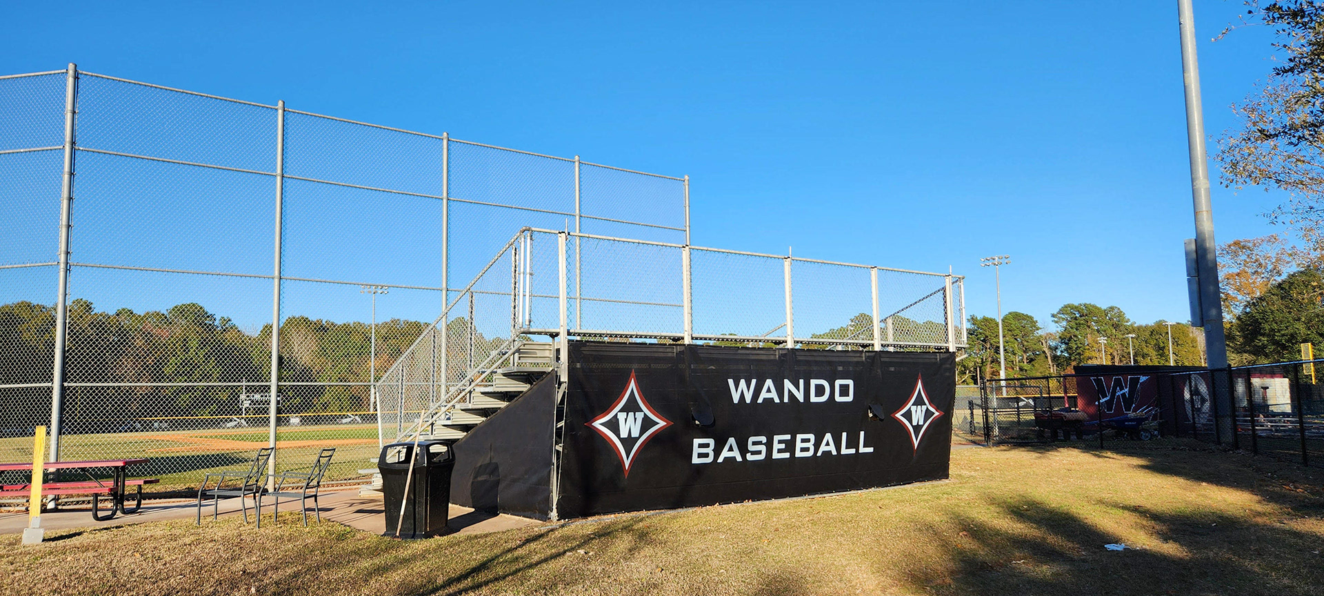 A black windscreen surrounds metal bleachers near a baseball field. The windscreen features the text 'Wando Baseball' in white, with the 'W' logos on both sides. Each 'W' logo consists of a large navy four-point star outlined in white and red, with a white 'W' in the center of the star.