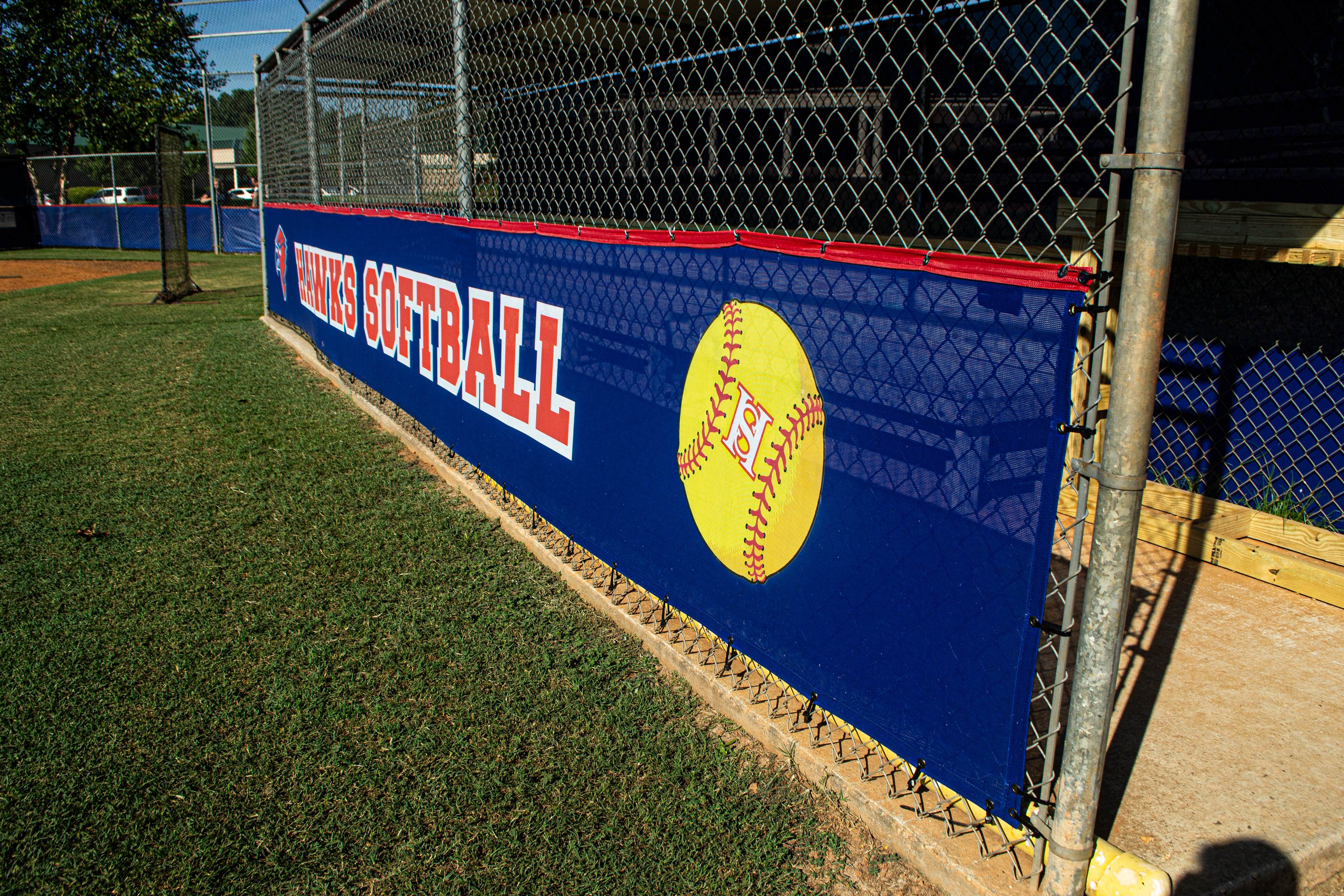 A baseball dugout with a fenced front, featuring a branded windscreen with red binding attached to the bottom half. Viewed from the front right side, the windscreen displays, from left to right: a red cardinal head logo, the text 'HAWKS SOFTBALL' in red outlined in white, and a yellow baseball with red lacing with a small white 'HS' logo outlined in red in the center.