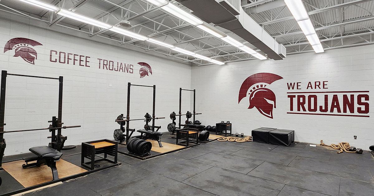 A weight room with a painted white brick wall featuring adhesive vinyl decals. On the left wall, two Trojan helmet silhouette decals face each other, with the words 'COFFEE TROJANS' positioned in the middle. On the right wall, there is a Trojan helmet silhouette with the words 'WE ARE TROJANS' next to it.