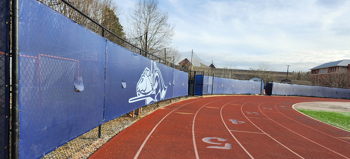 Side shot of navy blue fence windscreen panels showing their curvature alongside a football field, featuring a white outline of a knight riding a horse in a jousting position.