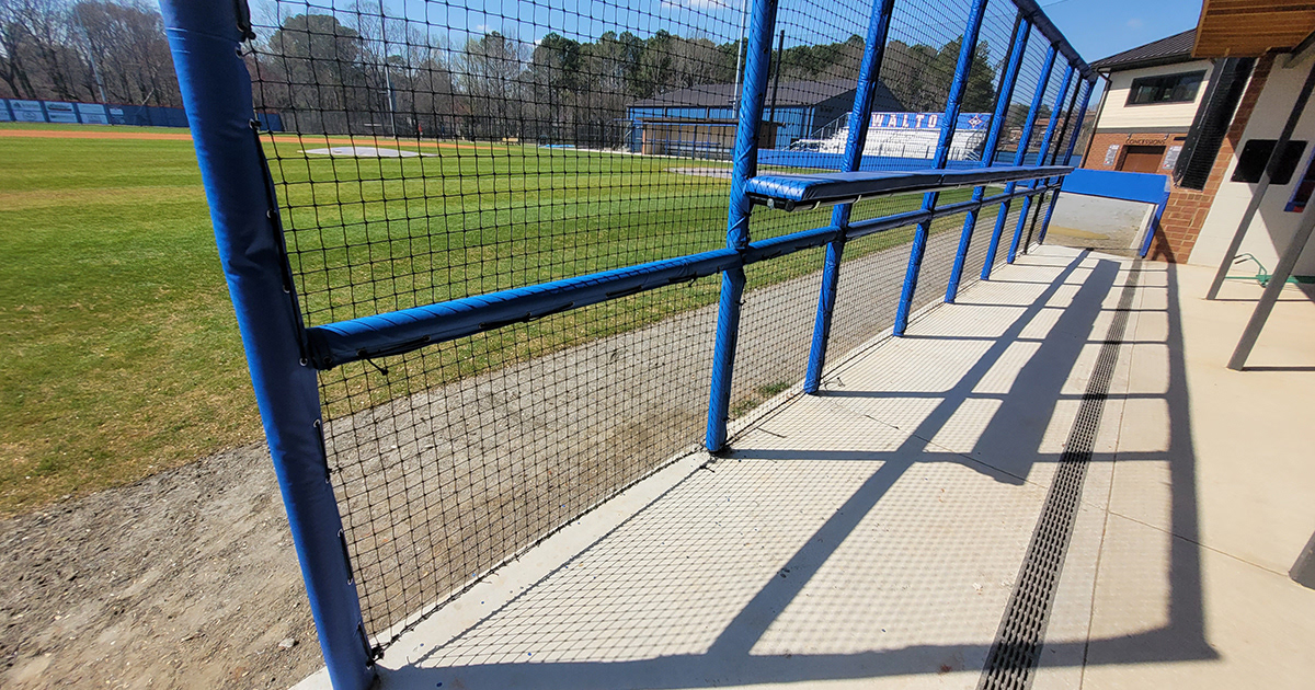 This image shows the round rail foam padding covered with blue sports vinyl and black sports netting in front of a baseball dugout barrier, captured from the back for a different perspective. 
