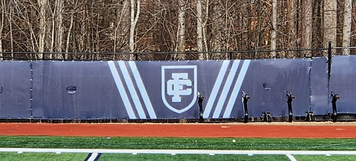 Close-up of a navy blue fence windscreen panel alongside a football field, featuring a white 'FC' monogram logo flanked by three vertical white stripes that angle inward at the bottom and outward at the top.