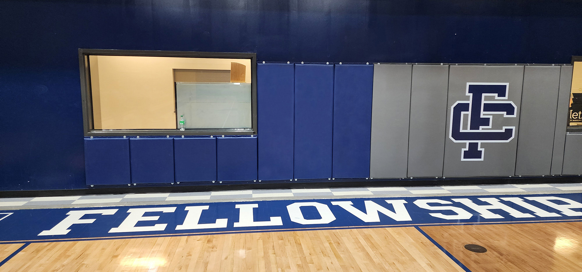 Thirteen vinyl-covered backstop pads in a gym, attached to a wall below and beside a set of windows. The left pads are blue, and the right pads are gray with a navy 'FC' monogram logo printed in navy blue and outlined in white.
